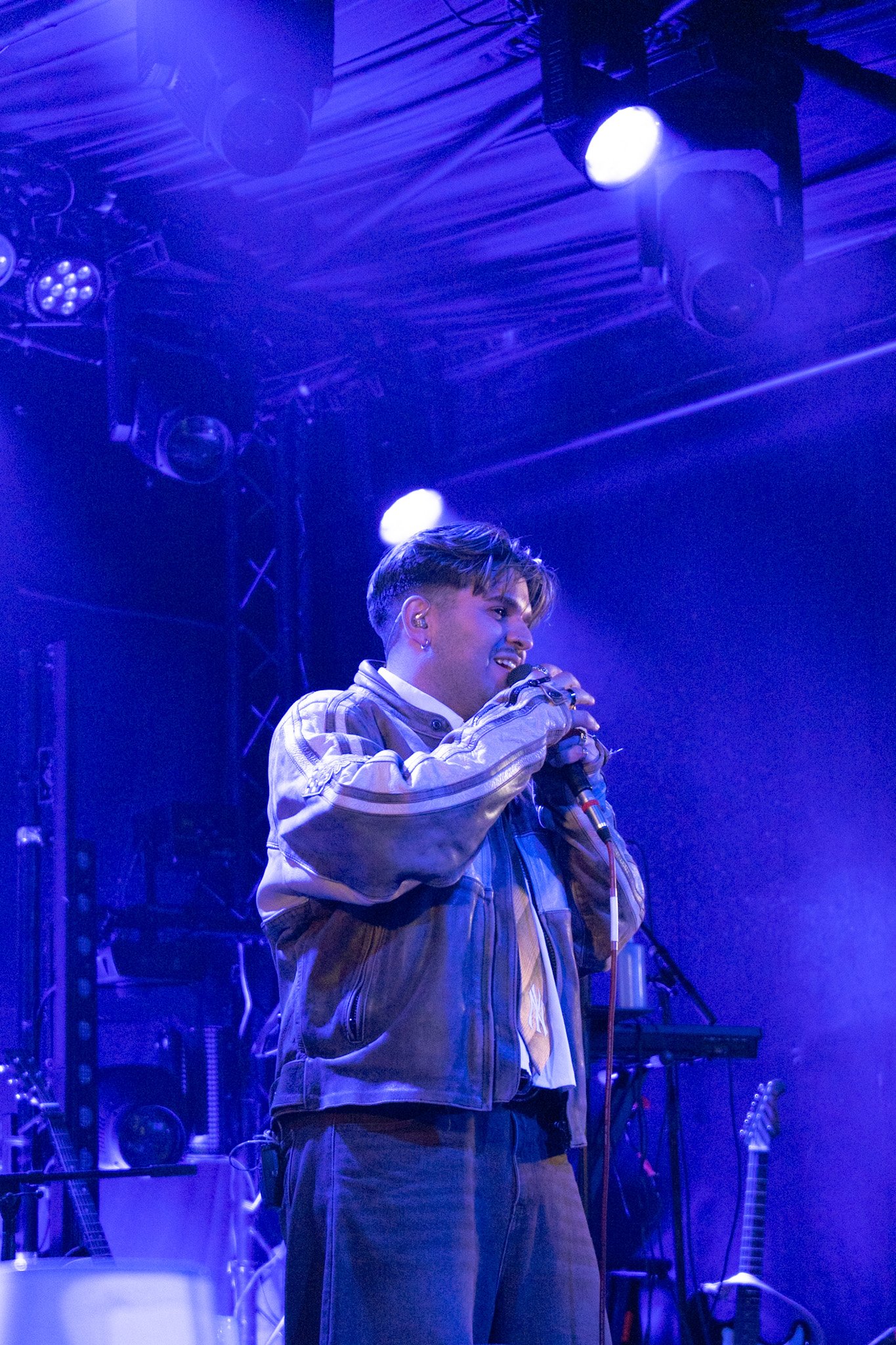 A young man singing into a microphone on stage with blue lighting, wearing a beige checkered jacket and dark pants, with musical instruments and stage equipment in the background.
