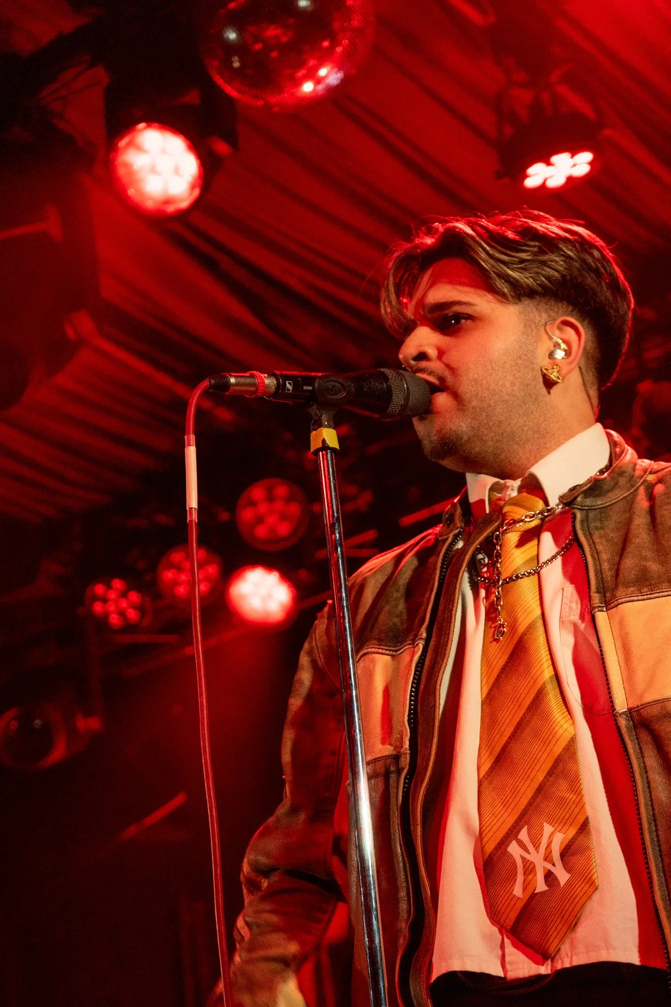 A man singing into a microphone during a live performance in a red-lit room.
