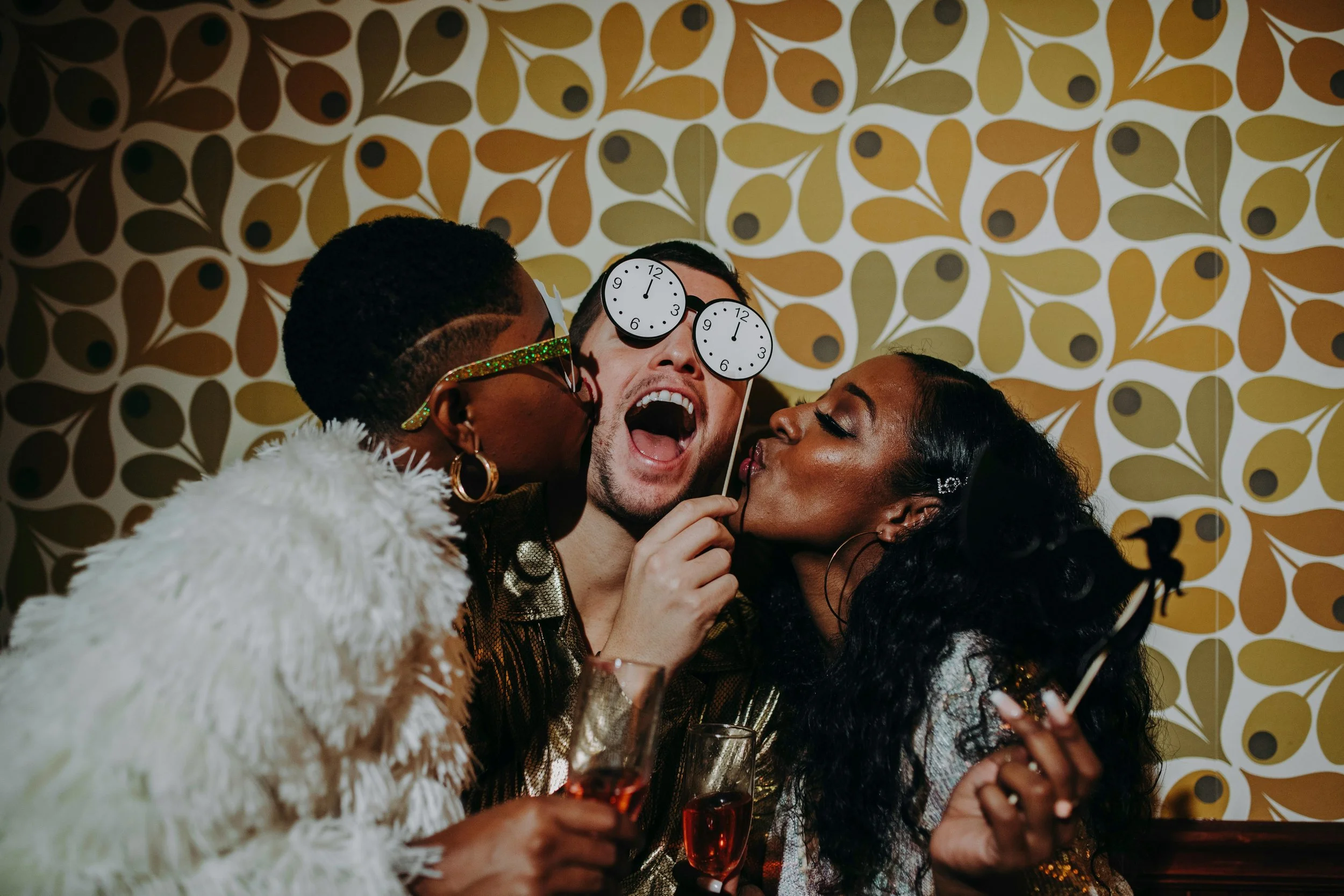 Three friends celebrating at a party in front of a digital photo booth photo, two women and a man, with two clocks on sticks and drinks, in front of a retro patterned wallpaper.