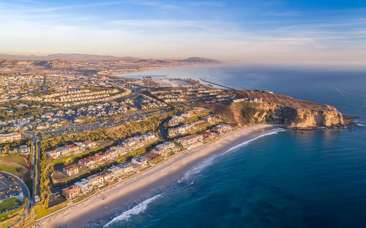 Aerial view of a coastal city with residential houses, a beach, and cliffs along the shoreline, with the ocean extending into the horizon under a partly cloudy sky.