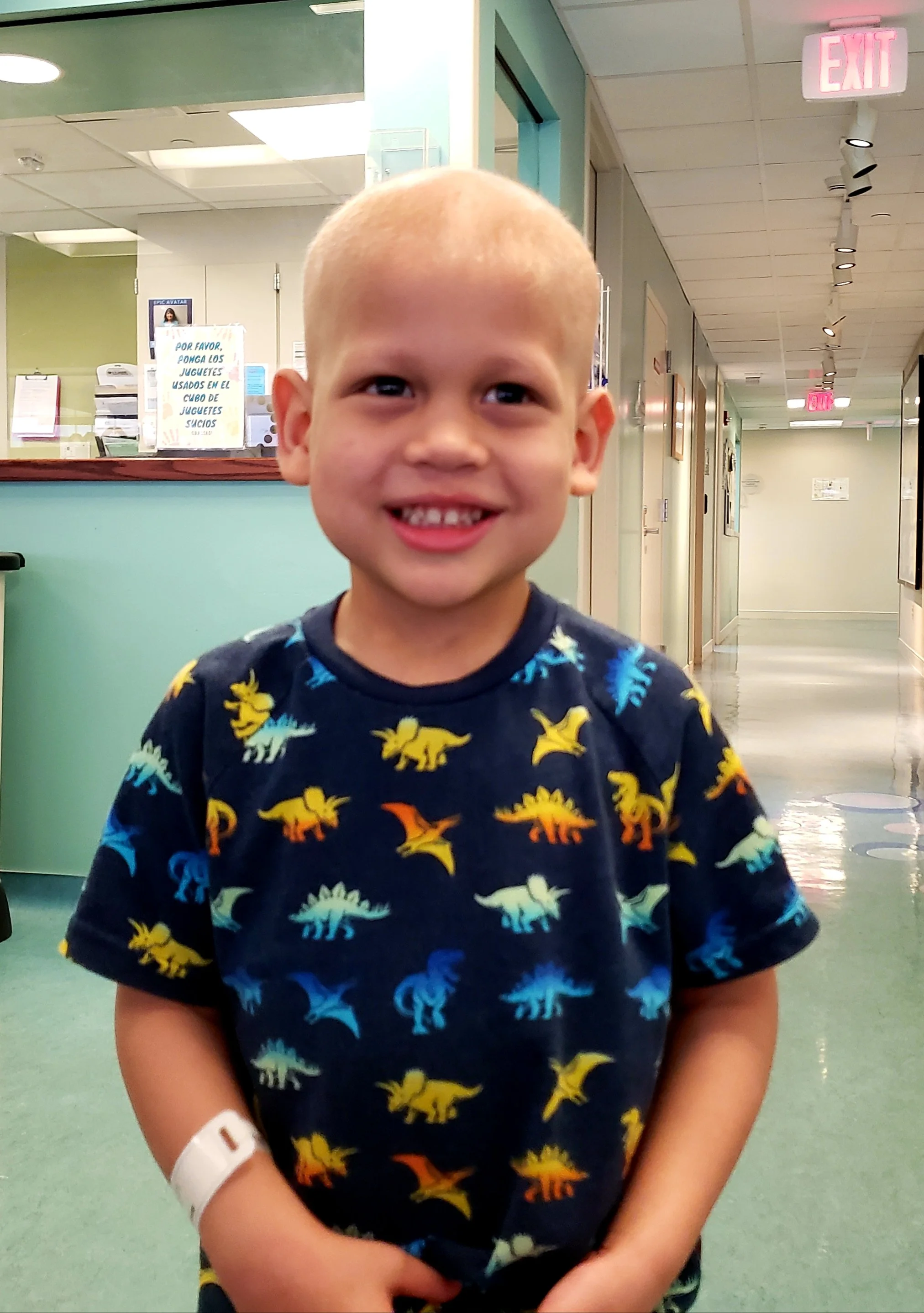 A young boy with a shaved head, smiling, standing in a hallway of a healthcare facility or hospital. He's wearing a dark t-shirt with colorful dinosaur prints and has a white hospital wristband on his left arm.