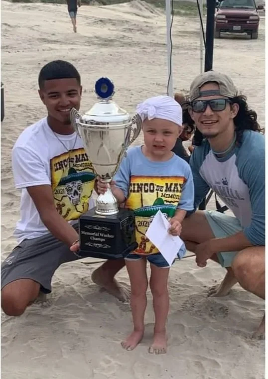 Three people on a beach holding a large trophy. The two adults are smiling, and the child in the middle is holding a piece of paper. All three are wearing T-shirts with a colorful design that says "El Cinco de Mayo" and a year, 2023.