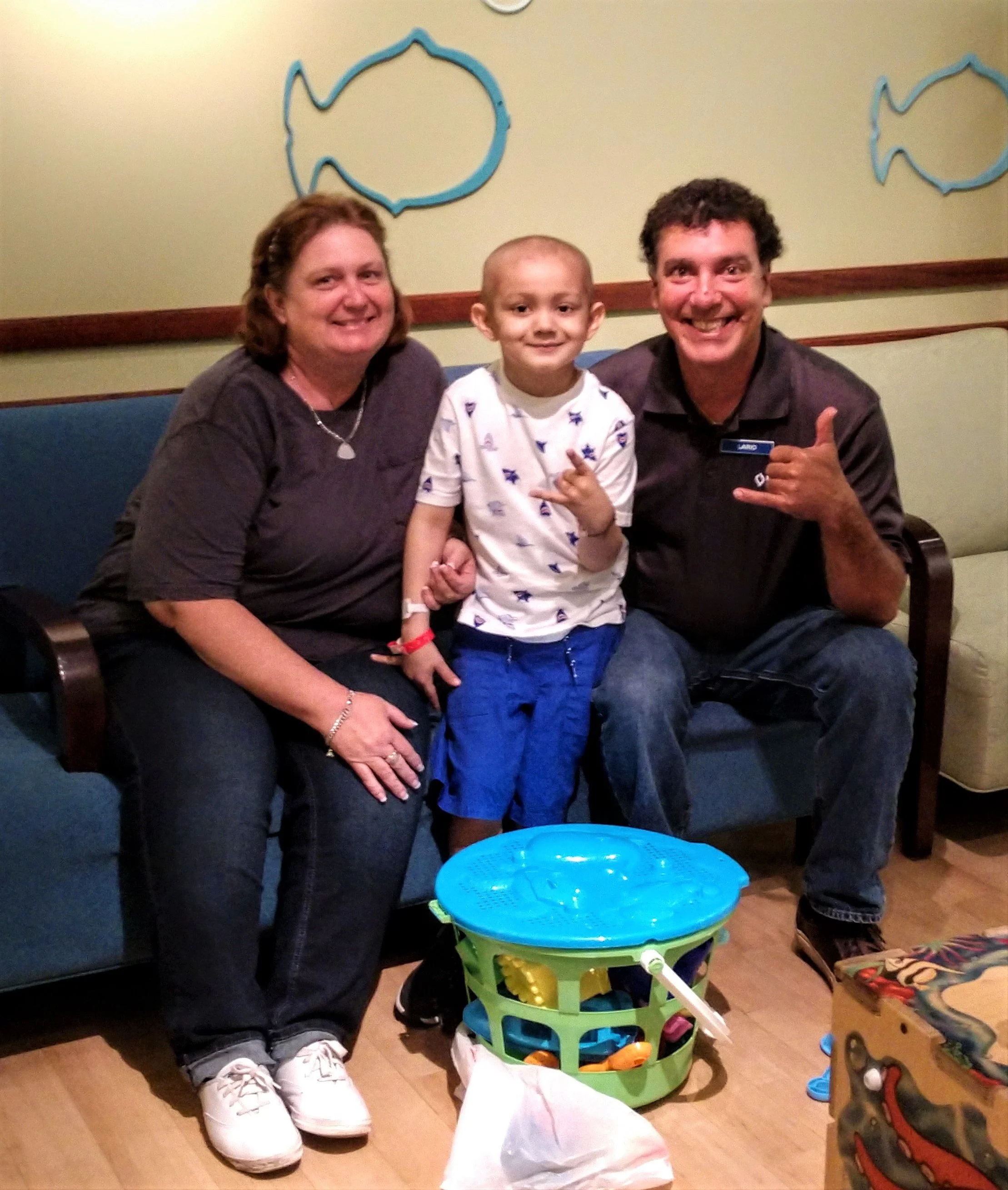Three people sitting on a bench, a woman on the left, a boy in the middle, and a man on the right, with toys on the floor and a wall with fish outlines behind them.