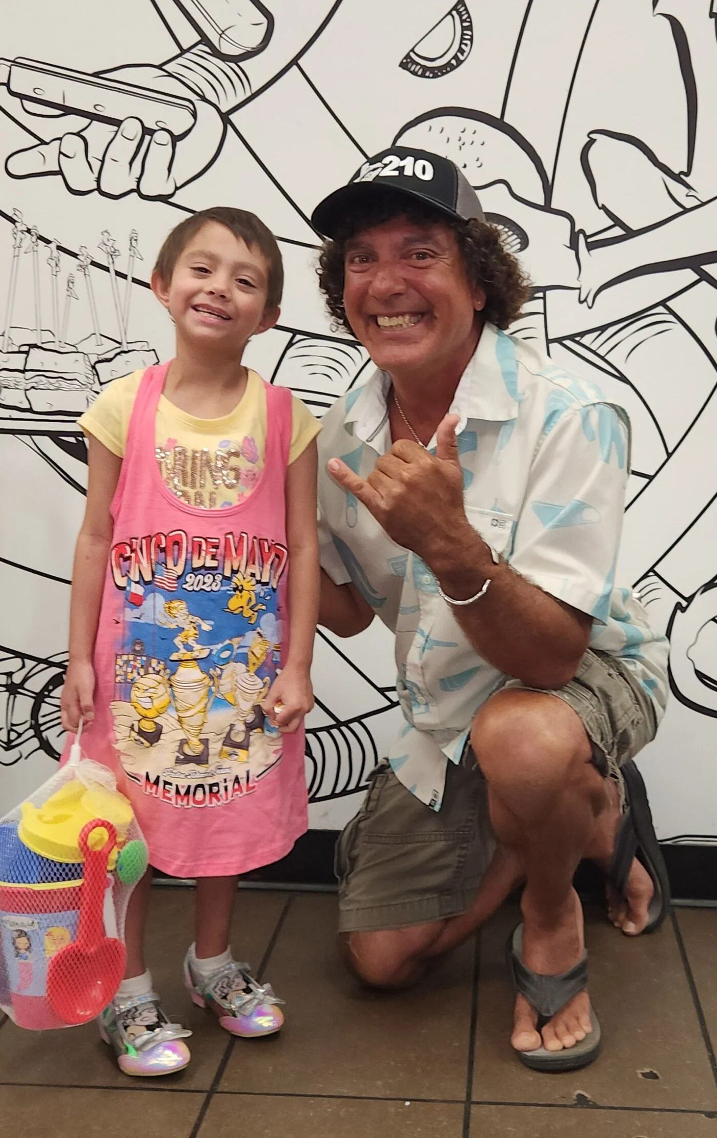 A man and a young girl smiling and posing in front of a black and white mural wall. The girl is holding a colorful toy bucket with a yellow lid, a green ball, and a red plastic shovel. The girl is wearing a pink apron with the words 'CND De Mayo 2023 Memorial' and cartoon characters, white socks, and shiny silver shoes. The man is wearing a Hawaiian shirt, khaki shorts, sandals, and a baseball cap, and is squatting on one knee with his right hand making a shaka sign.