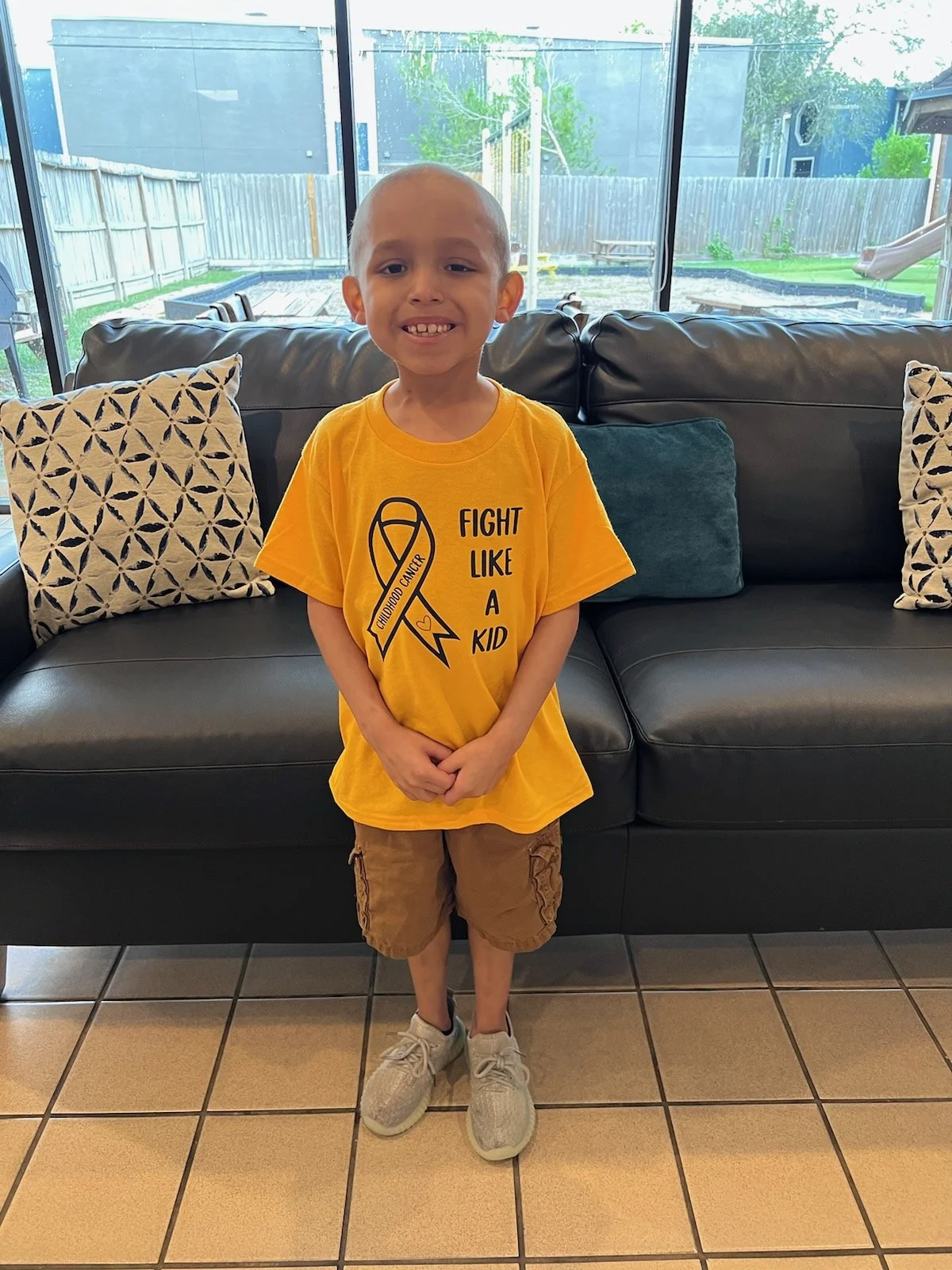 Young boy standing in front of a black leather sofa with patterned pillows, wearing a yellow T-shirt that says 'Fight Like a Kid' with a yellow ribbon symbol, brown shorts, and gray sneakers, smiling at the camera indoors with a view of a backyard through large windows.