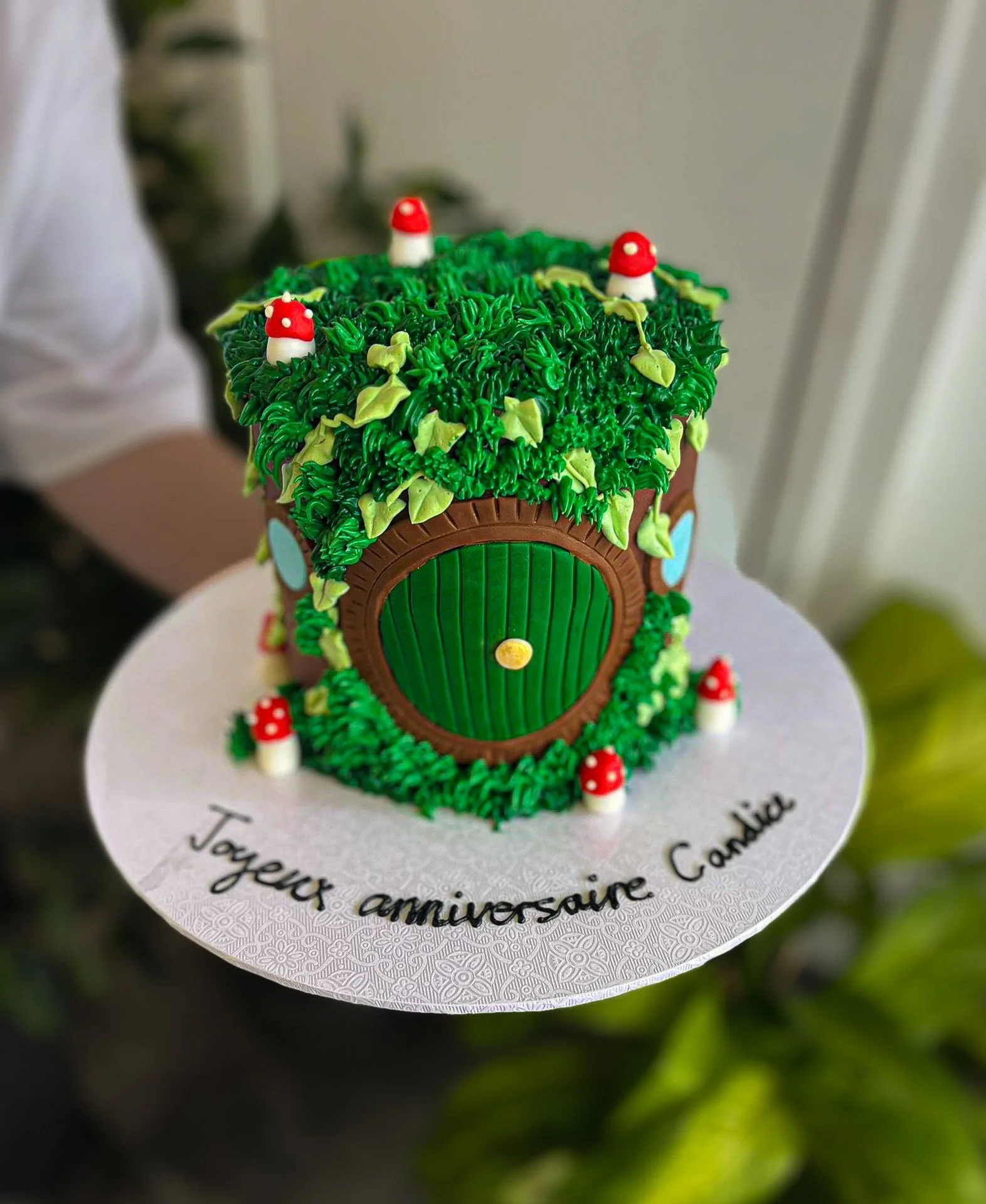 A birthday cake decorated to look like a small house with a green roof, brown walls, a green door, and red and white mushrooms, with a message in French on the base.