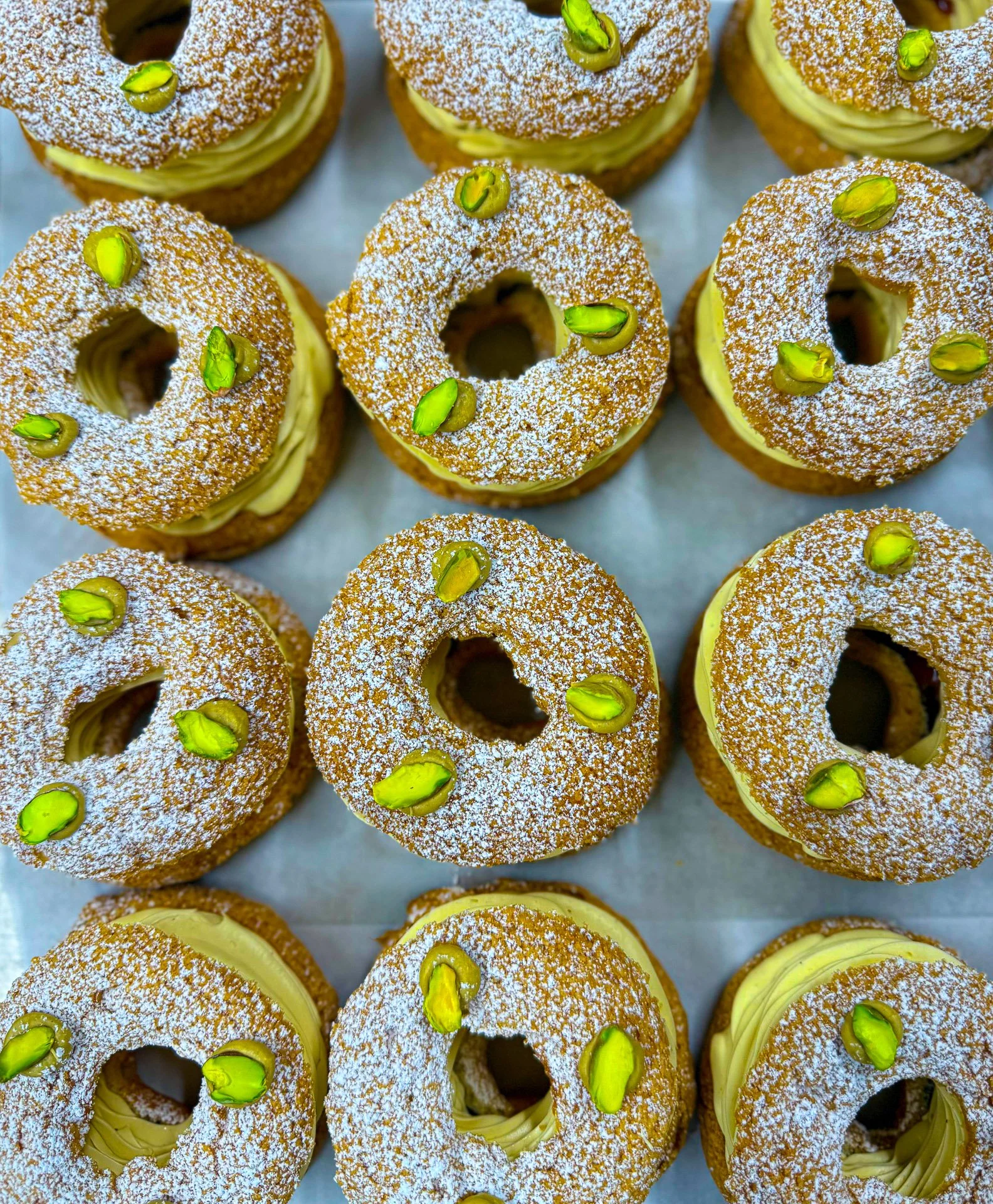 A tray of donuts topped with powdered sugar, green pistachio nuts, and filled with yellow custard or cream.