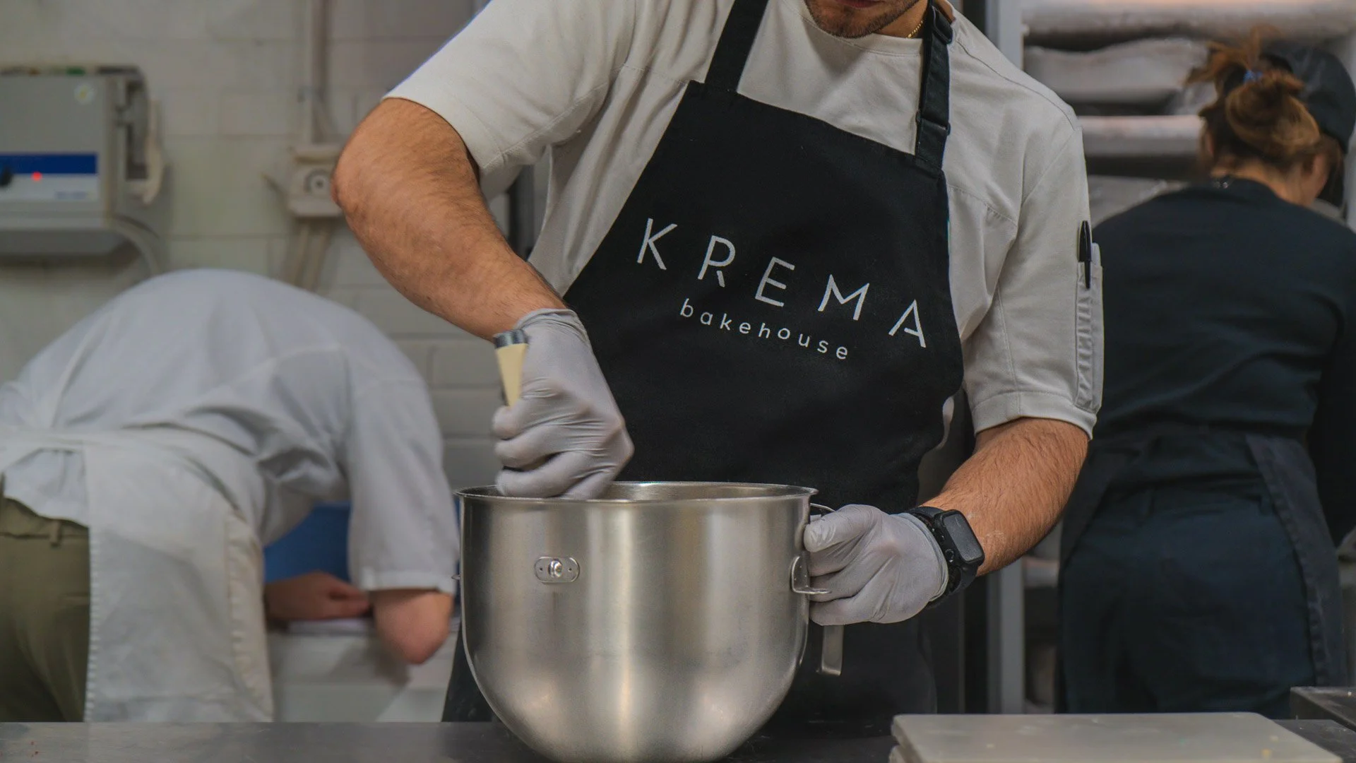 A person wearing a black apron labeled 'KREMA bakehouse' and gray gloves is stirring or mixing something in a large stainless-steel bowl in a commercial kitchen. Two other people are visible in the background.