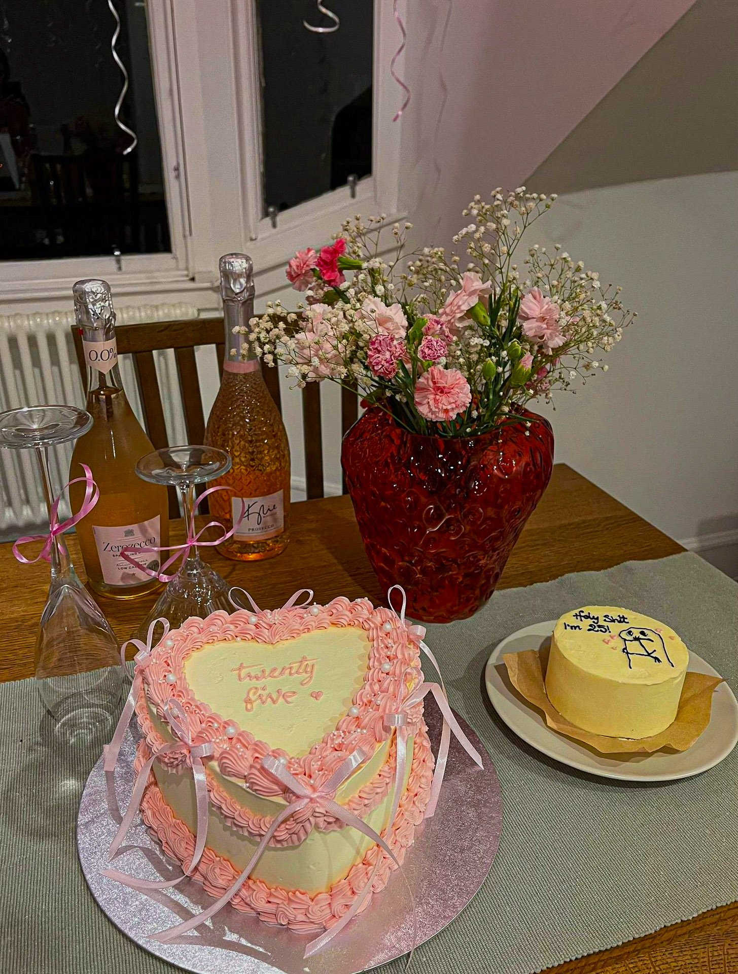 A table decorated with a pink heart-shaped cake with the words "twenty five" written on it, a yellow cake with a humorous message, two bottles of sparkling wine, two glasses, and a vase with pink and white flowers.