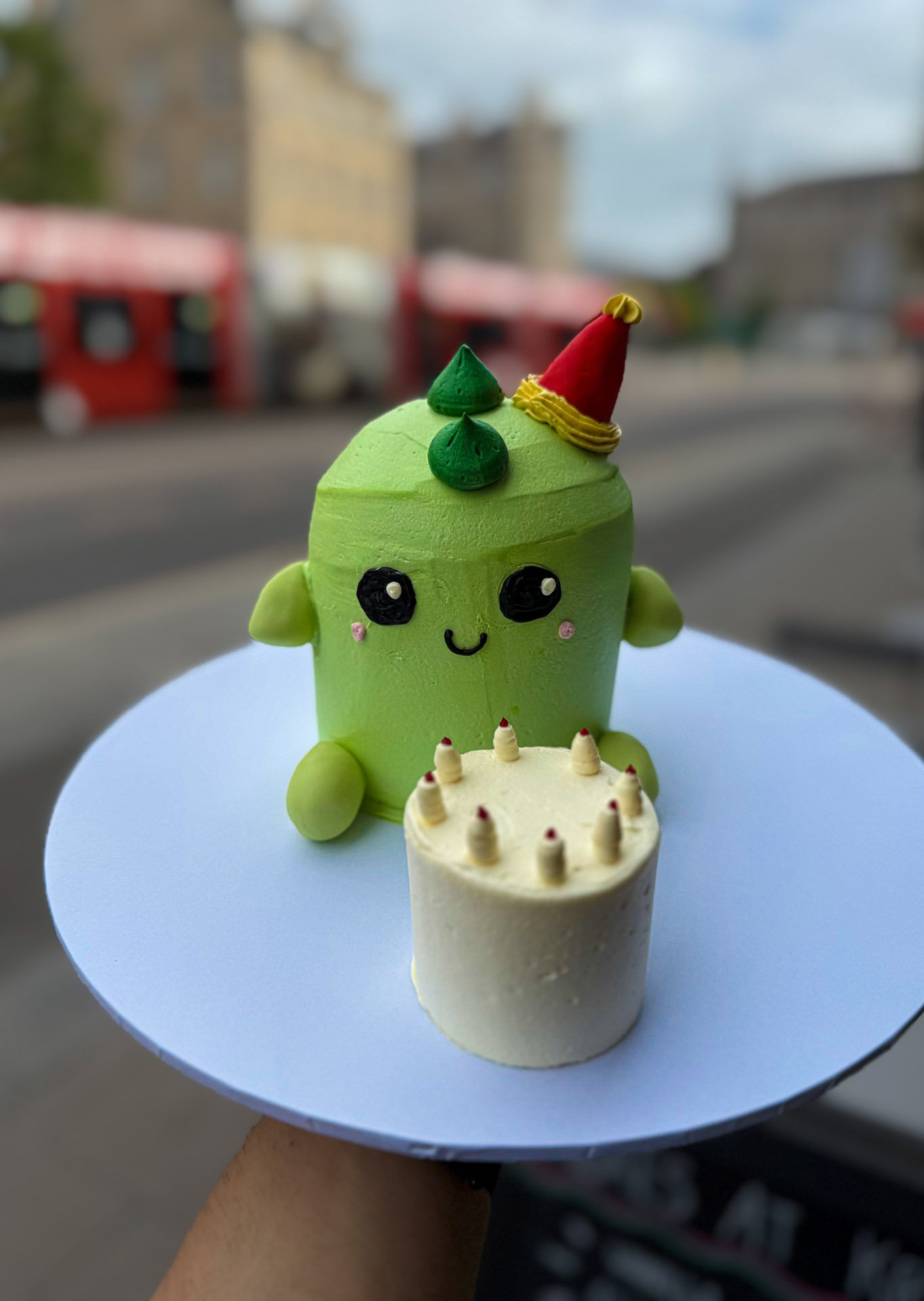 A small, cute cake shaped like a green character with a round body, big black eyes, a tiny smile, green ears, and decorated with a red party hat with yellow accents, sitting next to a smaller white birthday cake with seven lit candles on top on a whi