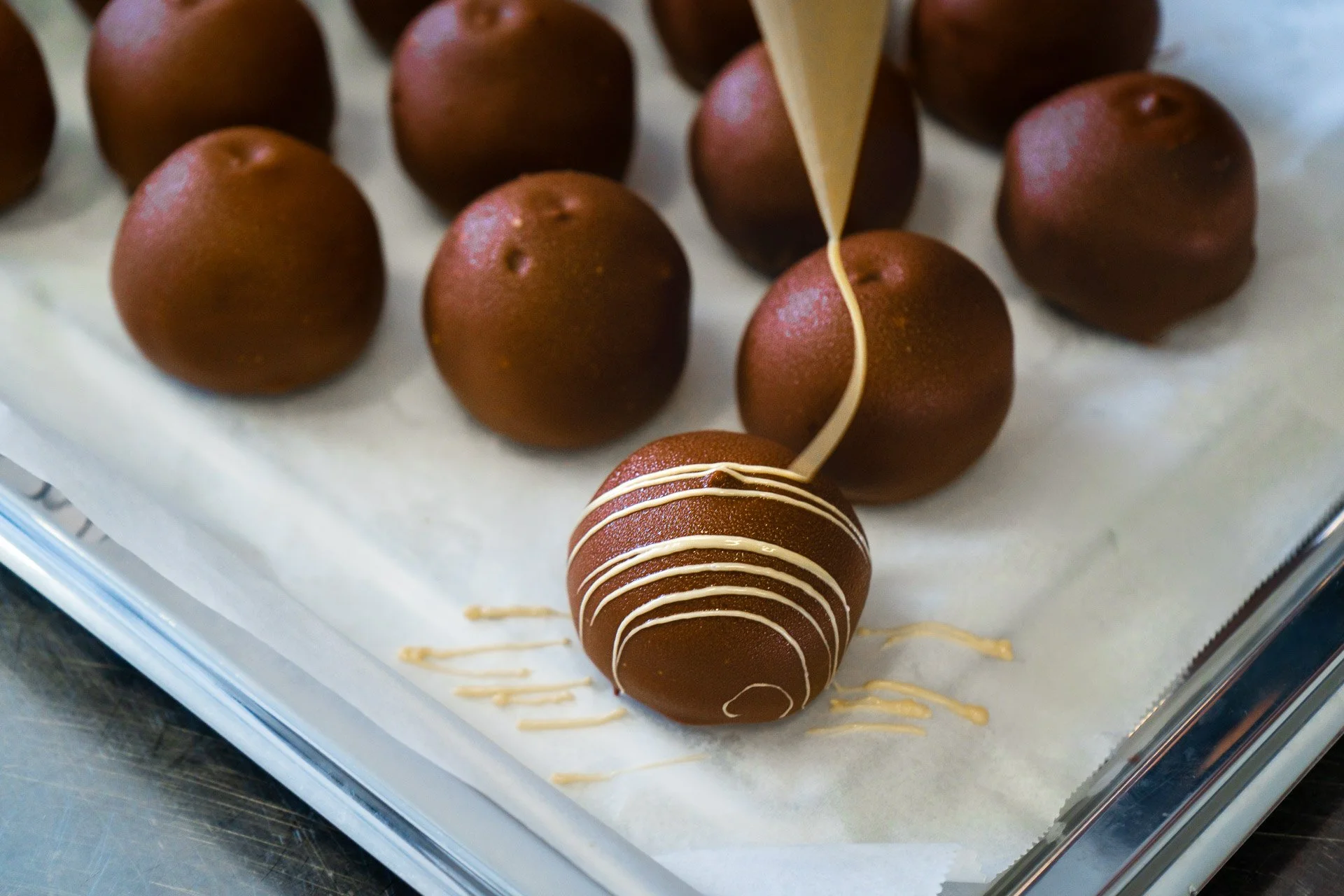Chocolate truffles being decorated with white chocolate drizzle on a baking sheet lined with parchment paper.
