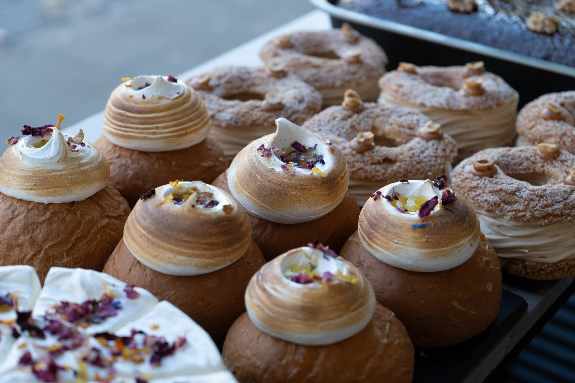 Assorted cream-filled pastries topped with edible flowers and garnishes on display at a bakery or market stall.