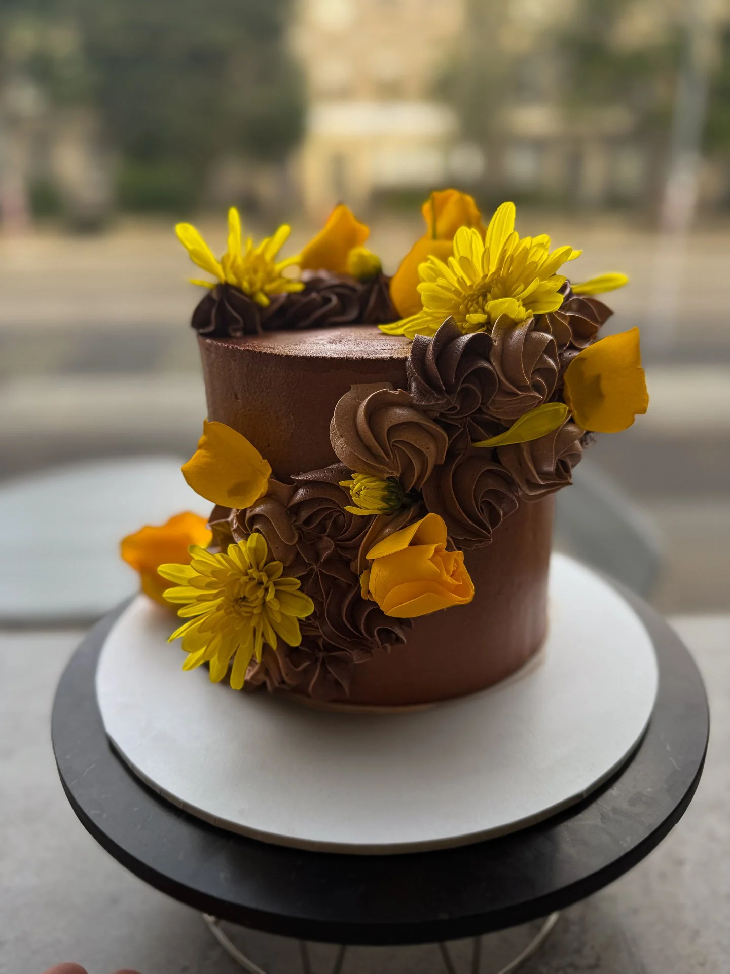 A chocolate cake decorated with yellow and orange flowers and chocolate frosting swirls, placed on a white cake board.
