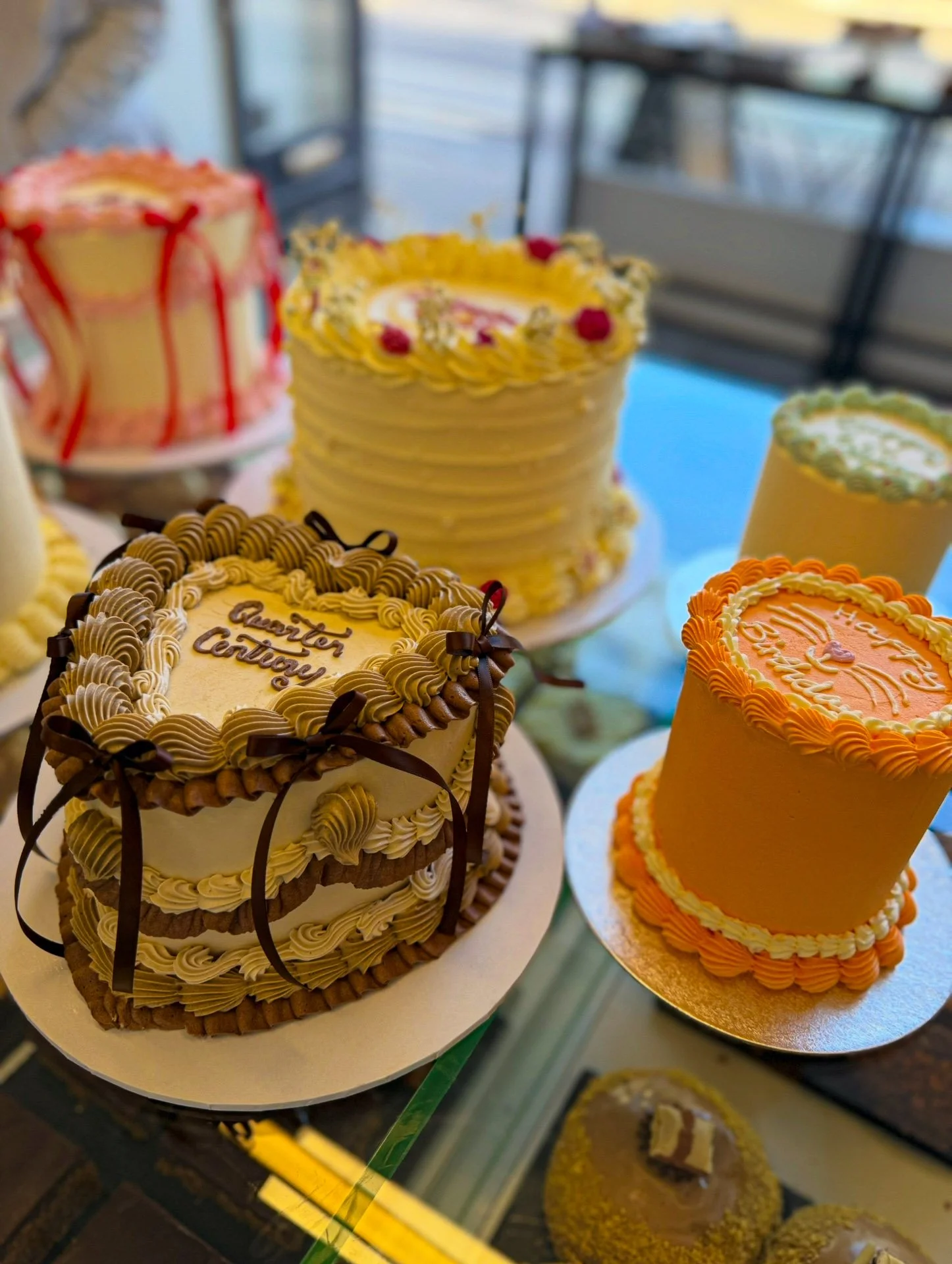 A display of various decorated cakes including a heart-shaped cake with chocolate ribbons and cream piping, a tall yellow cake with orange and white frosting, a large white cake with yellow frosting and red berries, and a pink cake with red drizzle.