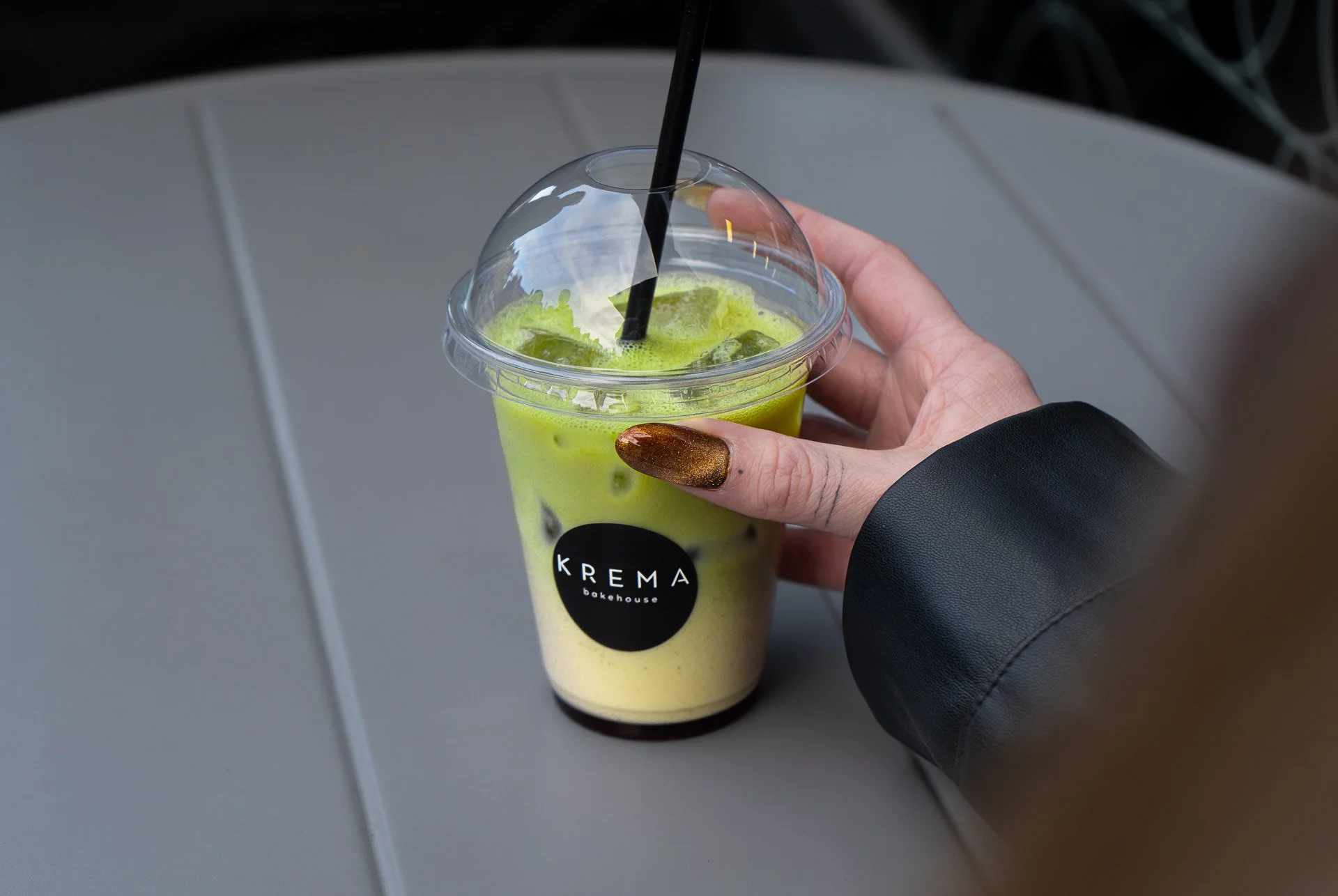 A hand with metallic brown nail polish holding a plastic cup with a black logo reading 'KREMA bakehouse', filled with green iced matcha latte and ice cubes, with a black straw.