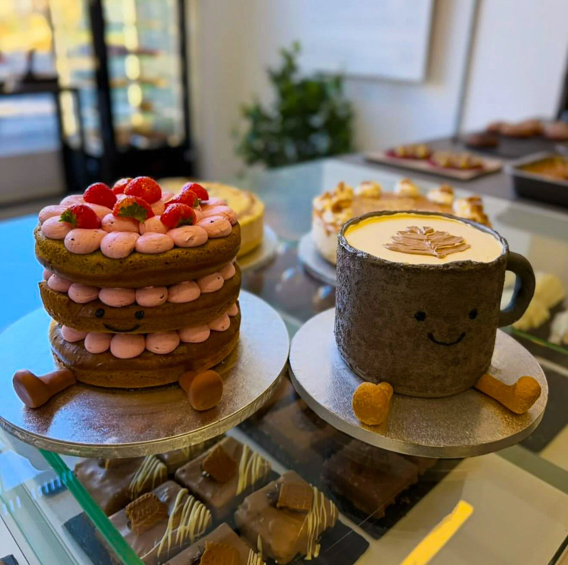 Decorative cake designed as a coffee mug with smiling face and two cookies as legs, and a stack of cookies with strawberries and pink cream, on a cake stand in a bakery display case.