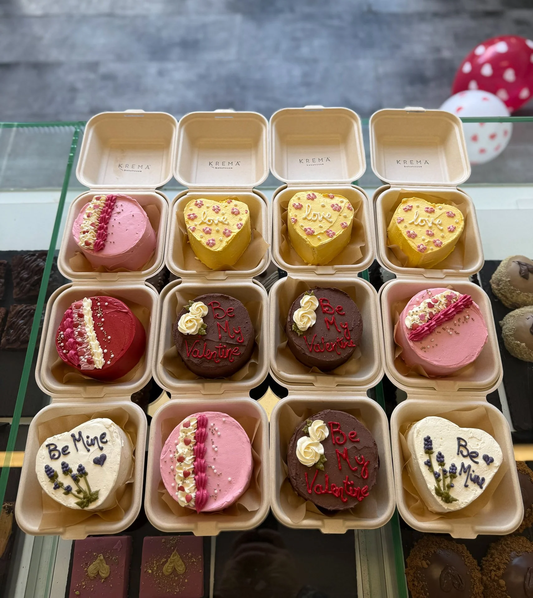 A display of assorted heart-shaped and round Valentine’s Day-themed desserts in beige boxes, decorated with words like 'love,' 'Be Mine,' and 'Be My Valentine,' topped with floral and ribbon decorations.