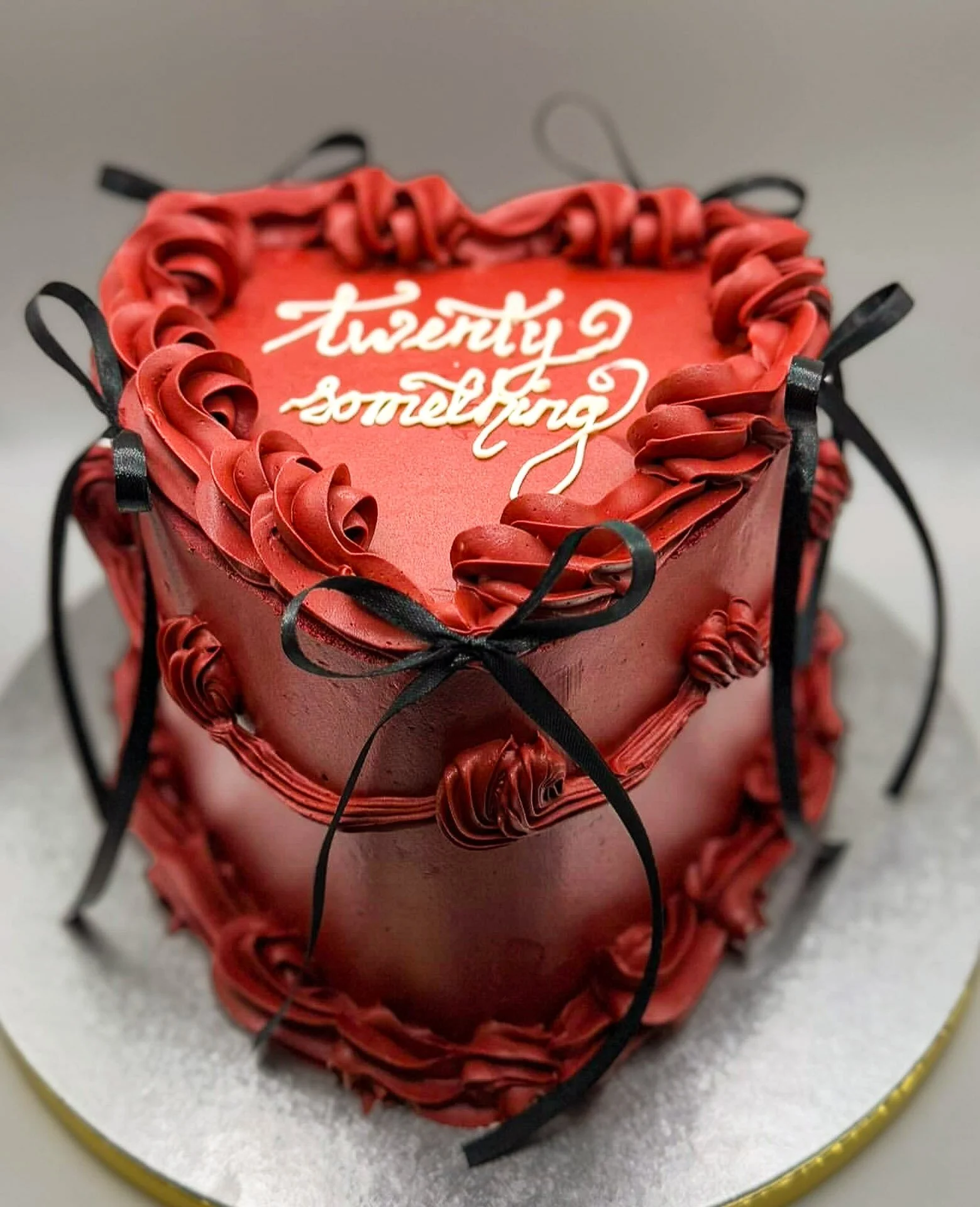A heart-shaped red cake decorated with red and black icing, with the words "twenty something" written on top in white icing.