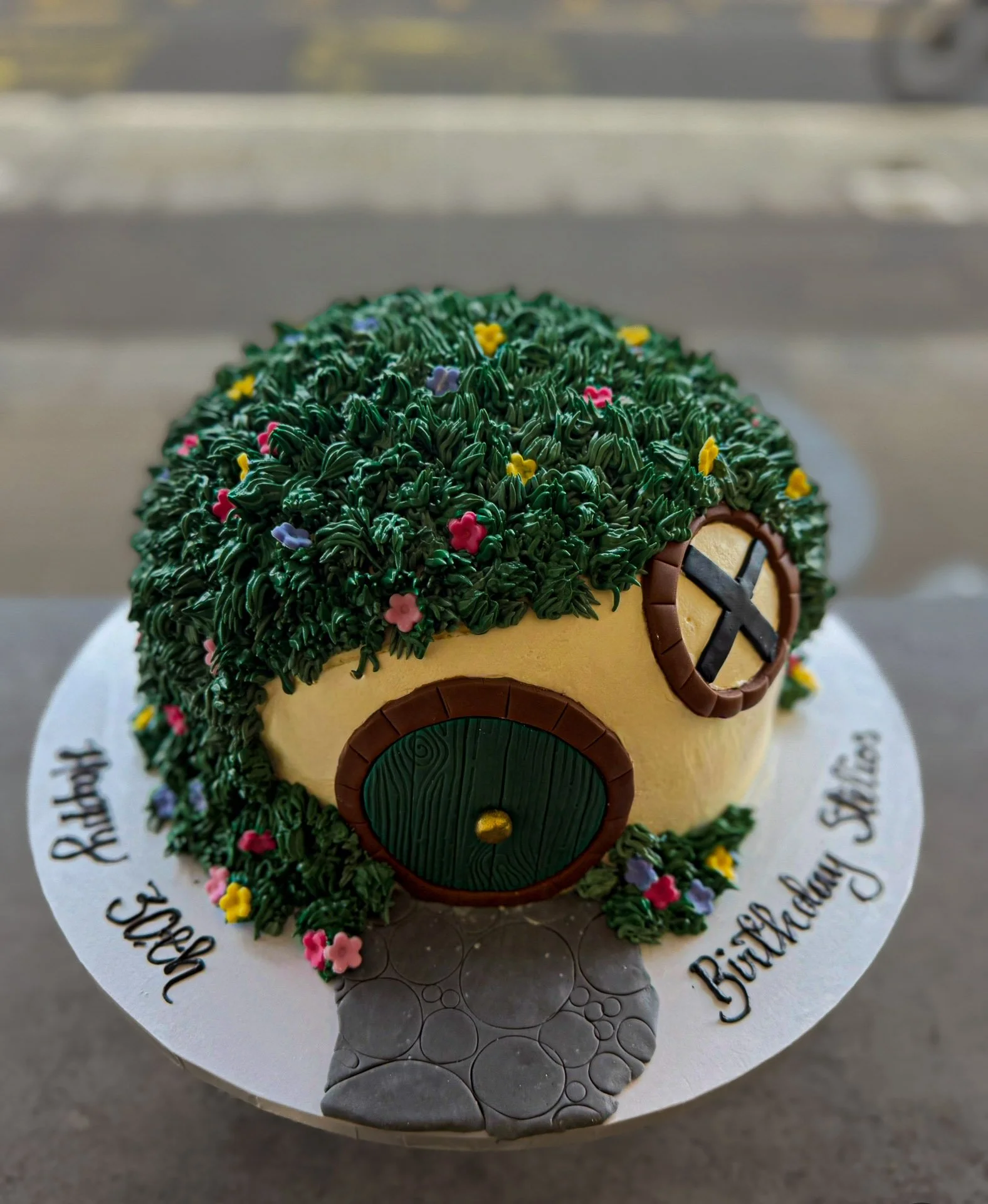 Round cake decorated to resemble a Hobbit house from The Lord of the Rings, with a green, grassy roof with small colorful flowers, a beige wall, a green door, and a circular window, on a white board with birthday message written around the edge.