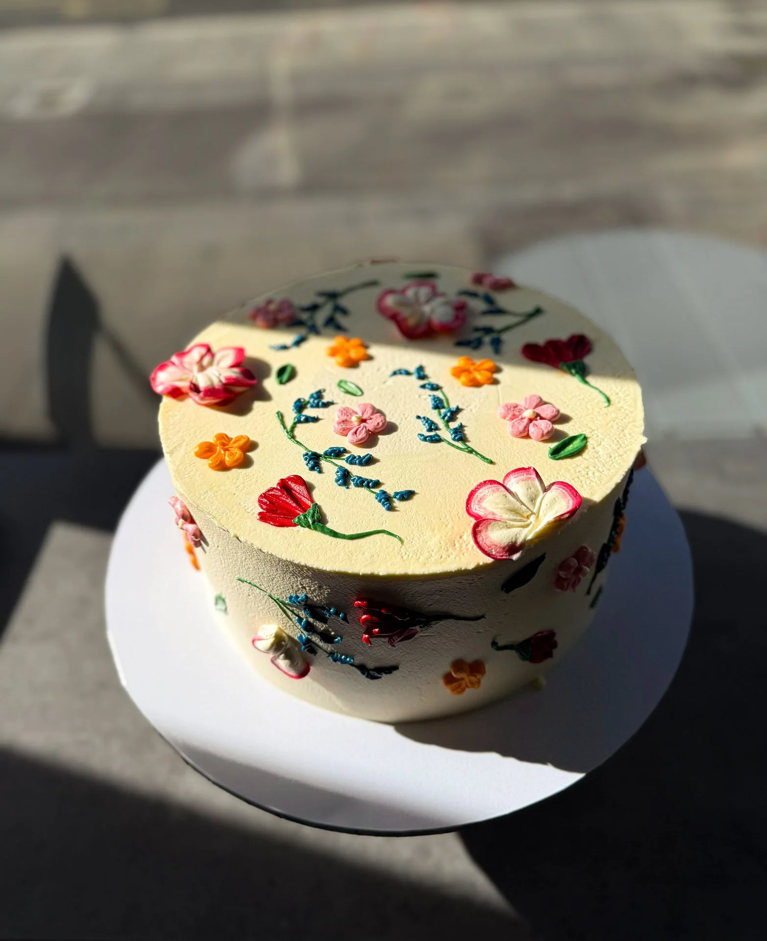A round white cake decorated with colorful floral designs and blue script on top, sitting on a white cake board.