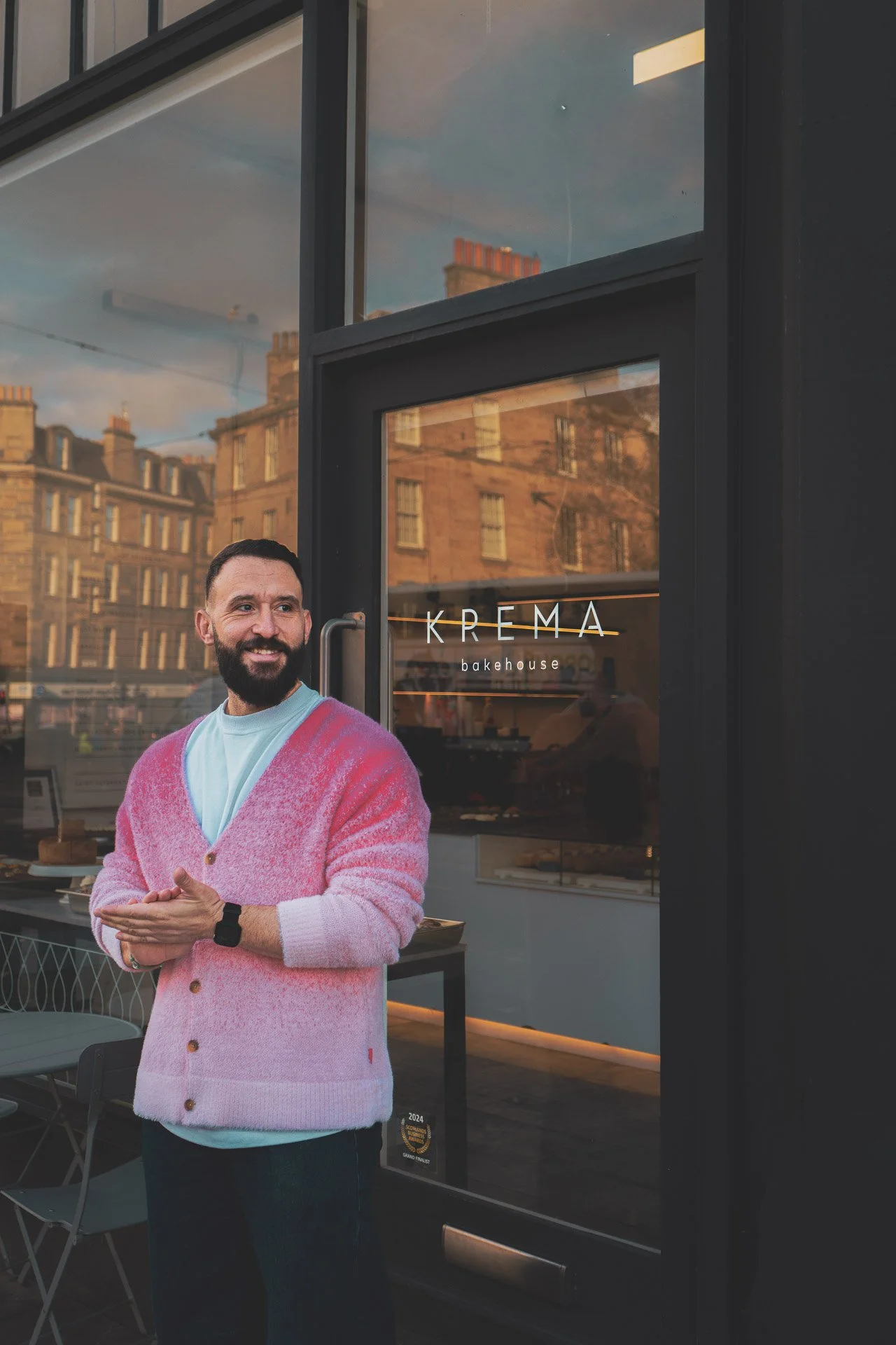 A man standing outside a bakery named "KREMA bakehouse" on a city street in the evening, wearing a pink and white gradient cardigan and smiling.