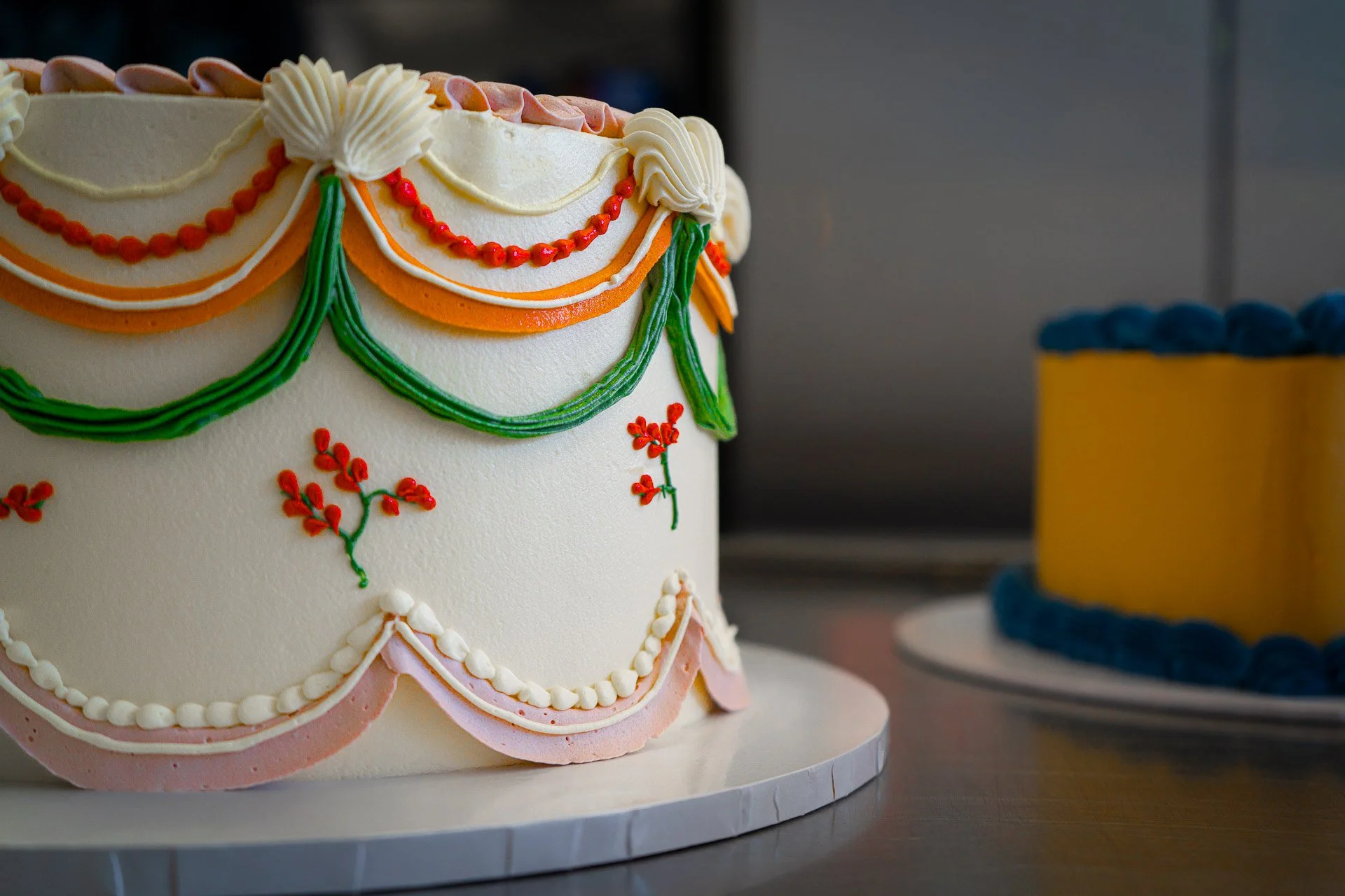 Decorated wedding or celebration cake with colorful icing drapes and floral details, partially visible in the foreground, and a second cake with a yellow and blue design in the background.