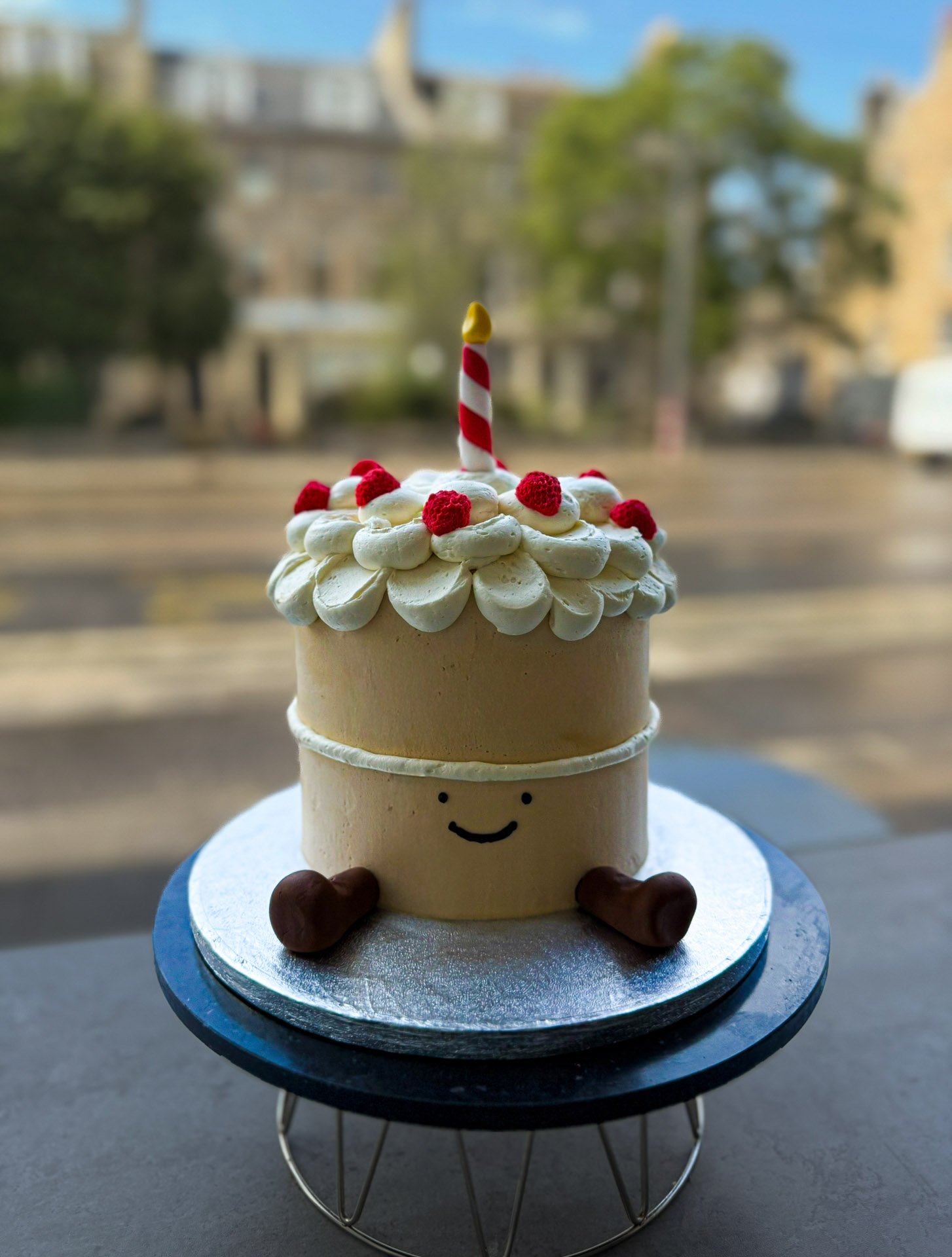 A cute cake with a smiling face, small brown legs, topped with white frosting, red raspberries, a candle, and decorated with piped frosting, sitting on a silver cake board.