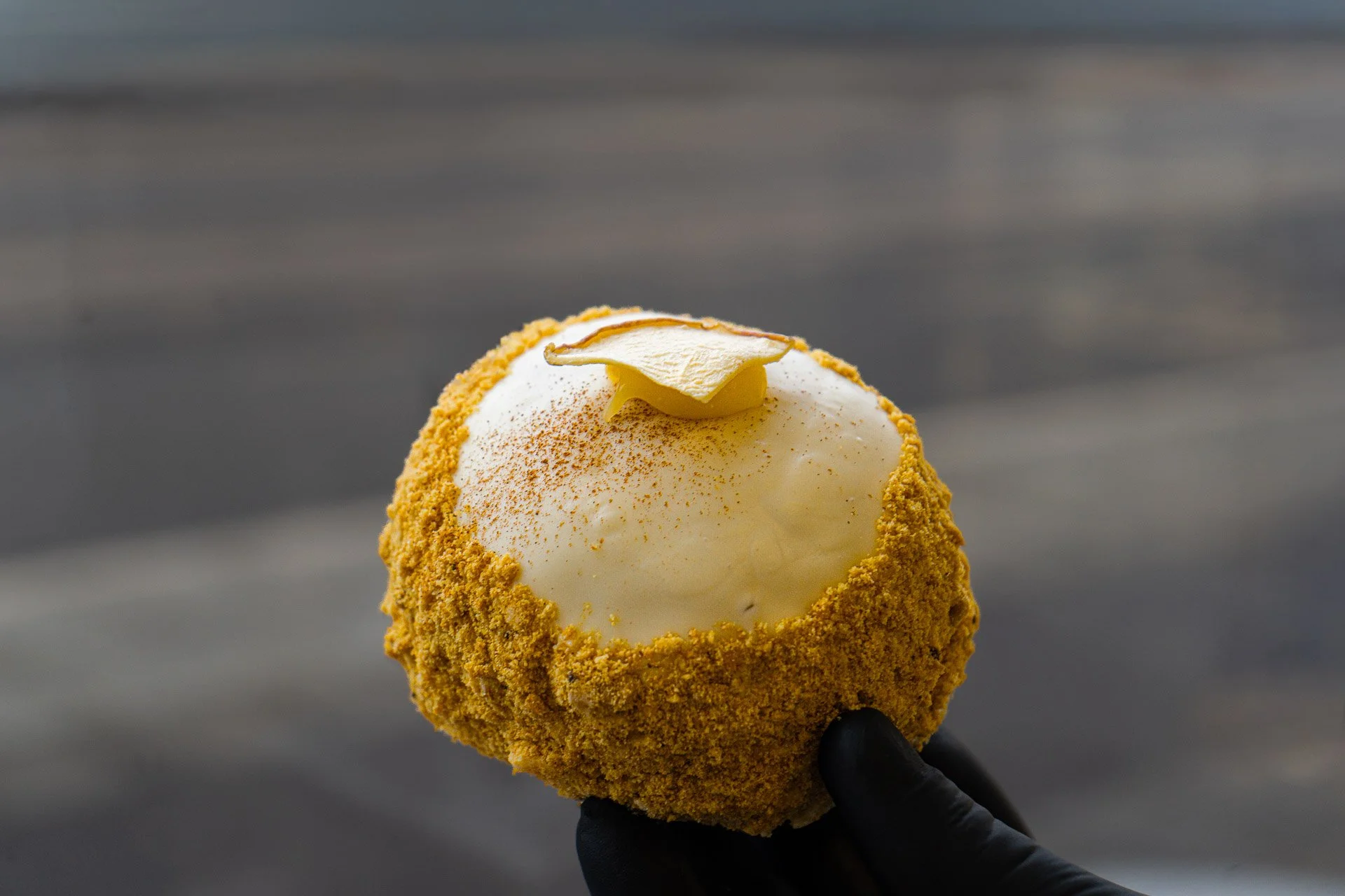 A close-up of a dessert consisting of a round, white scoop of ice cream or sorbet, topped with a thin slice of lemon and a dusting of cinnamon or nutmeg, with the scoop surrounded by a crumbly golden-brown coating, held by a hand wearing a black glove.