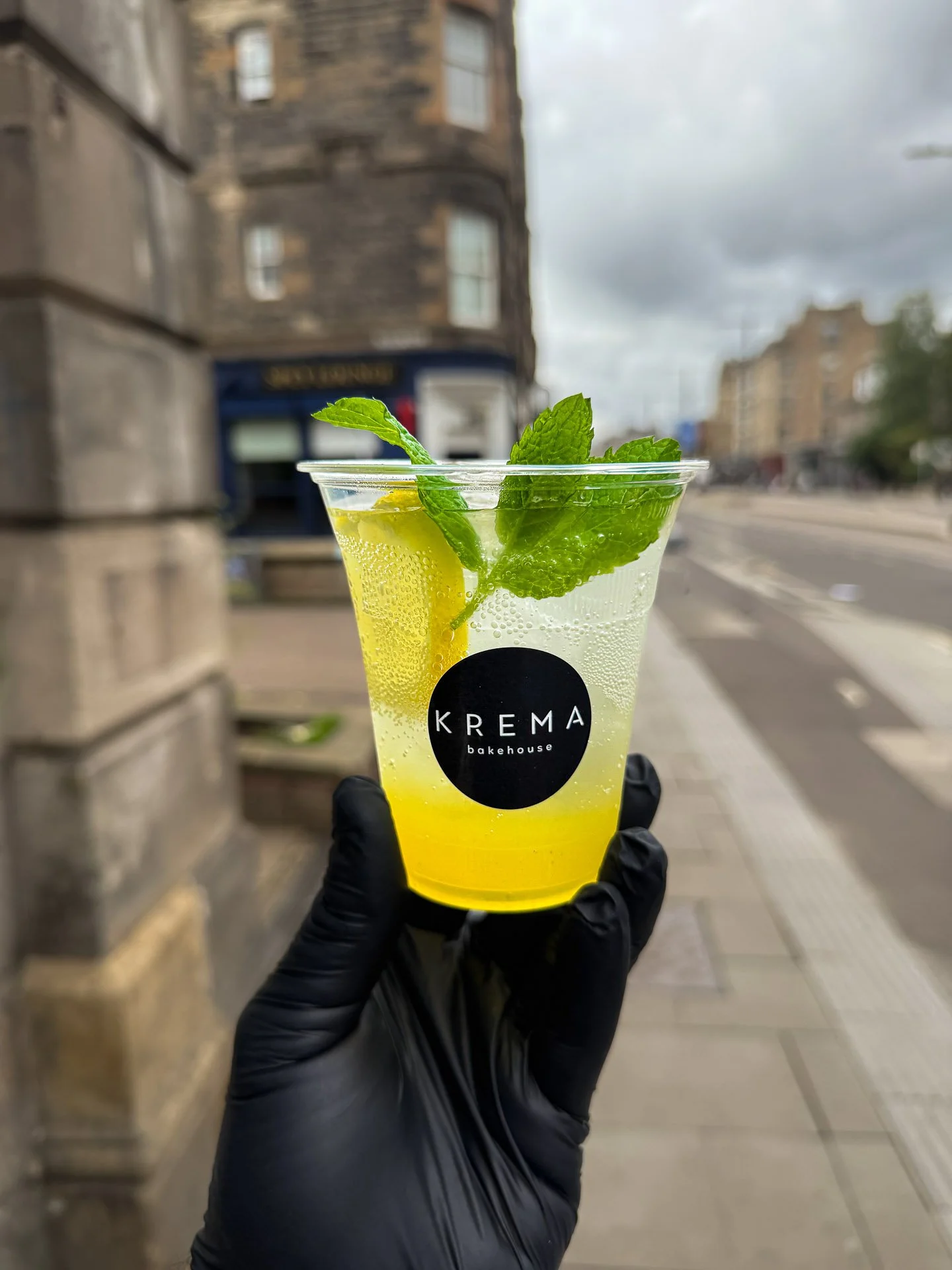 A hand wearing a black glove holds a clear plastic cup with a yellow-green beverage, garnished with lemon slice and fresh mint leaves, in an outdoor urban setting with cloudy sky and old stone buildings.