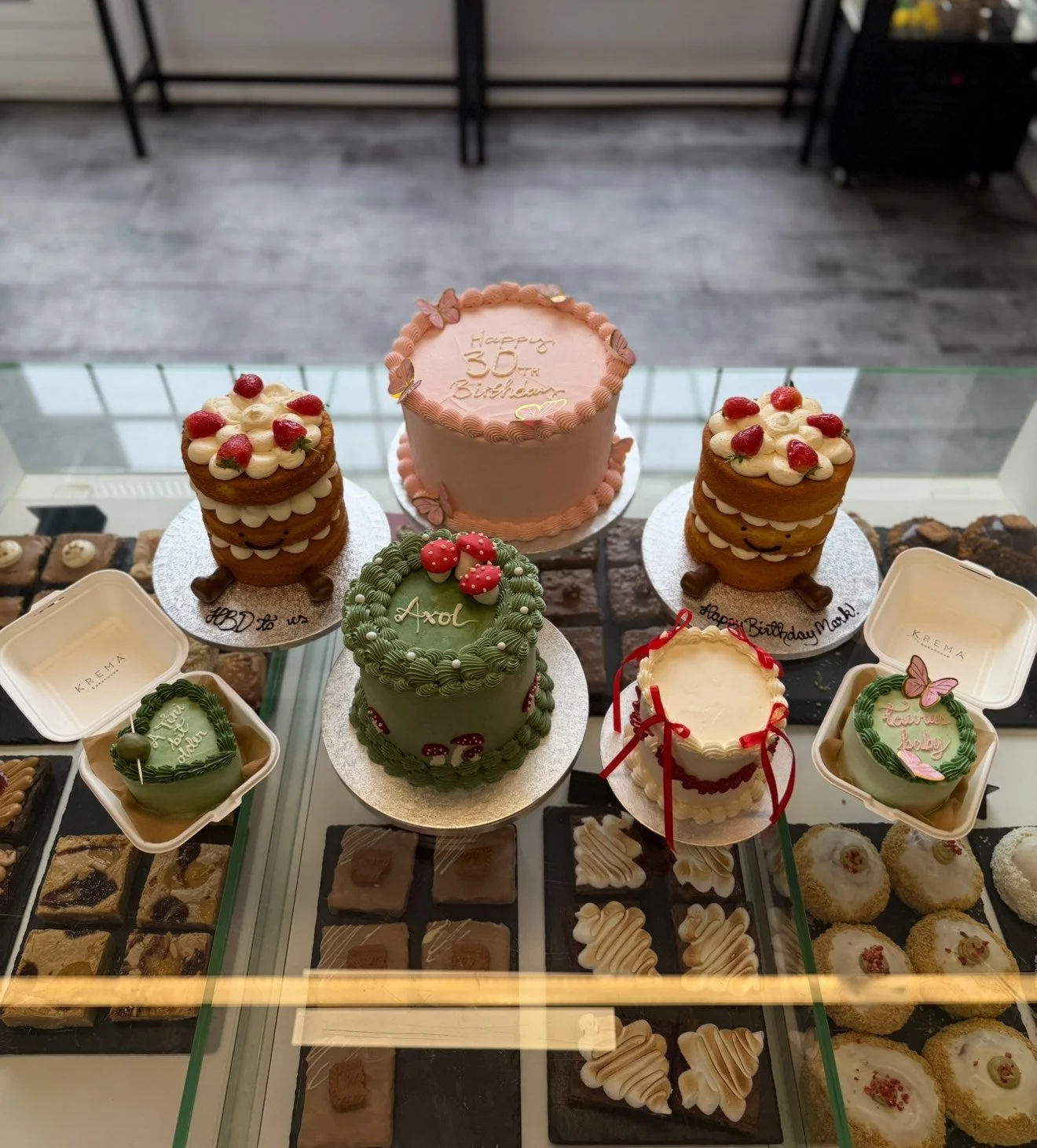 Assorted cakes and desserts on display. There is a pink birthday cake with "Happy 30th Birthday" written on it, two green decorated cakes with mushrooms and 