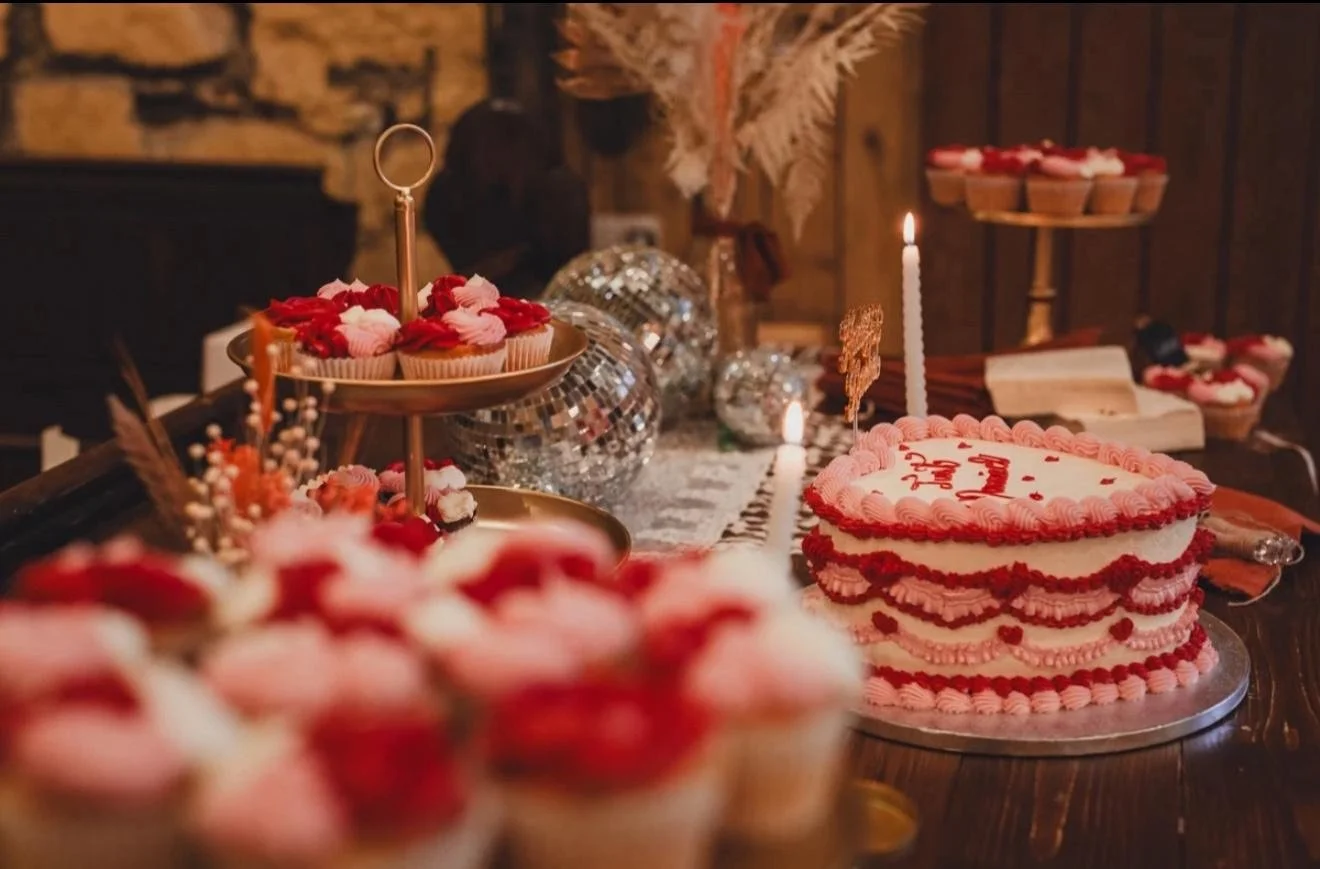 A heart-shaped cake decorated with pink and red icing, with a lit candle on top, on a table with assorted cupcakes, disco balls, and decorative items for a celebration.