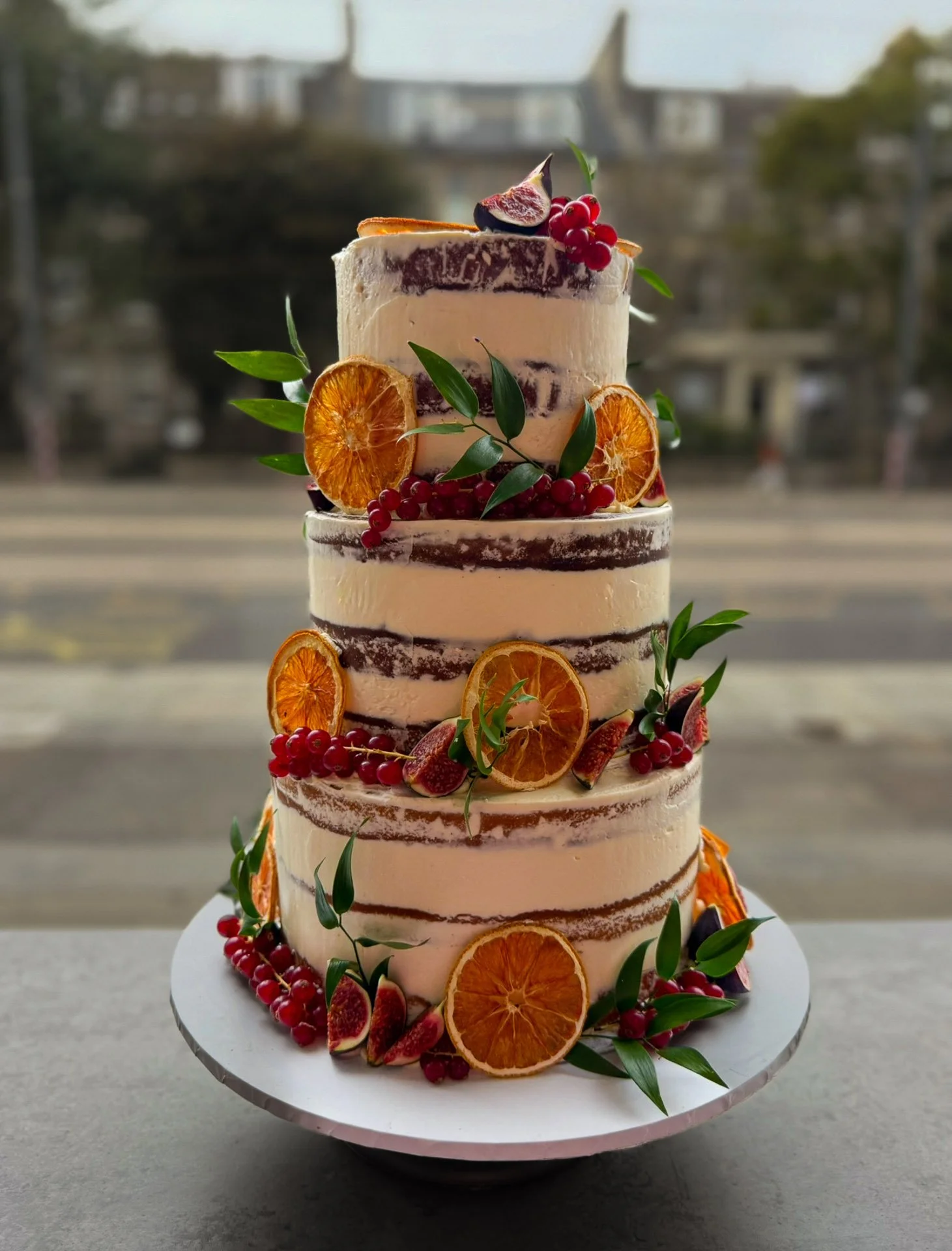 Three-tiered naked cake decorated with dried orange slices, red currants, and green leaves, set on a white cake stand with a cityscape background.