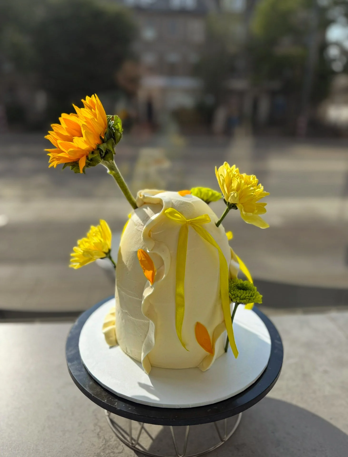 Cream-colored cake decorated with yellow and orange flowers and ribbons, placed on a stand in front of a window with a blurred city street background.