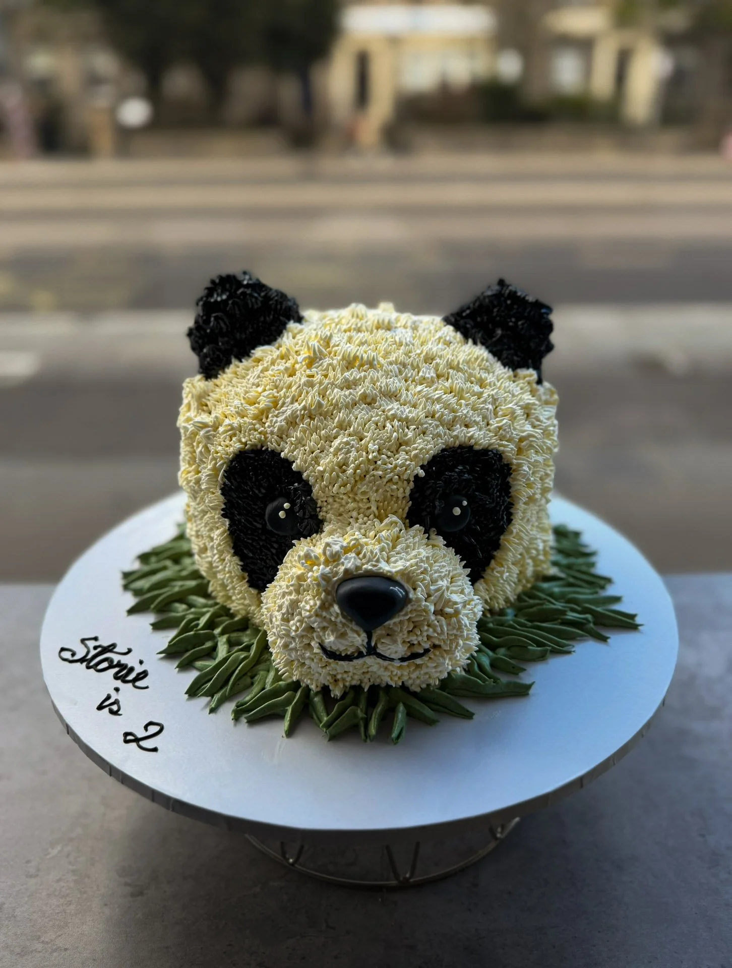 Panda-shaped cake with cream frosting, black fondant details, and a grassy base on a white cake board with the message 'Storie is 2' written on it.
