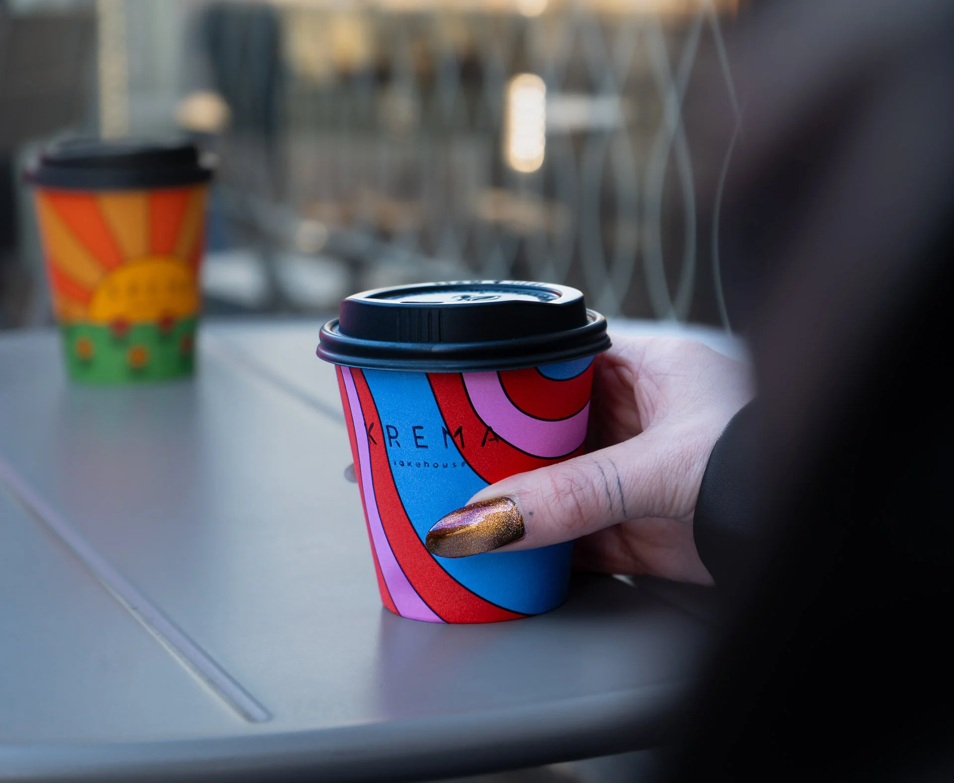 Person holding a colorful coffee cup with abstract swirling design, with another similar cup in the background on a gray surface near a metal railing.
