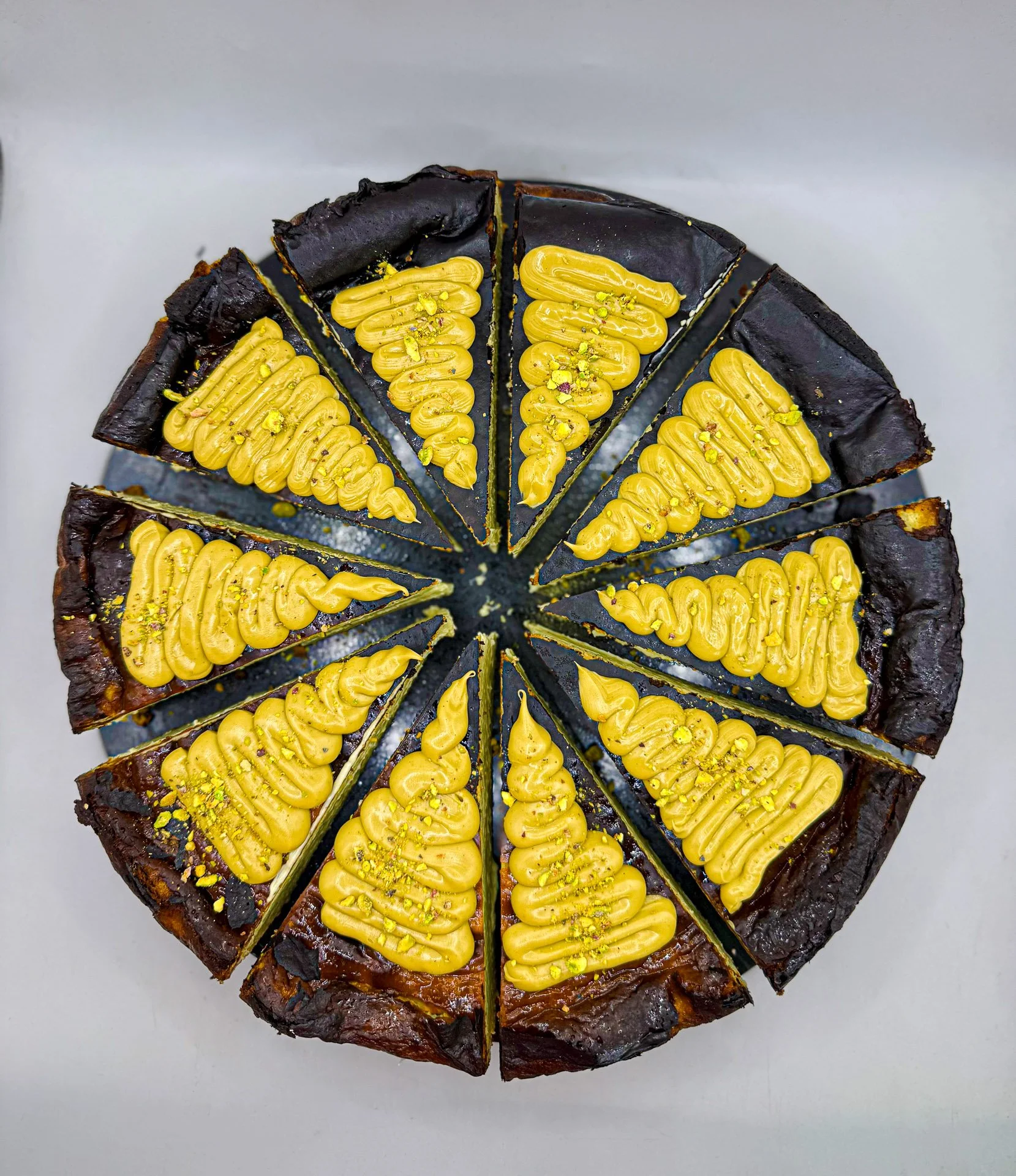 A round sliced dessert with a dark chocolate crust, topped with yellow piped frosting and sprinkled with crushed pistachios.