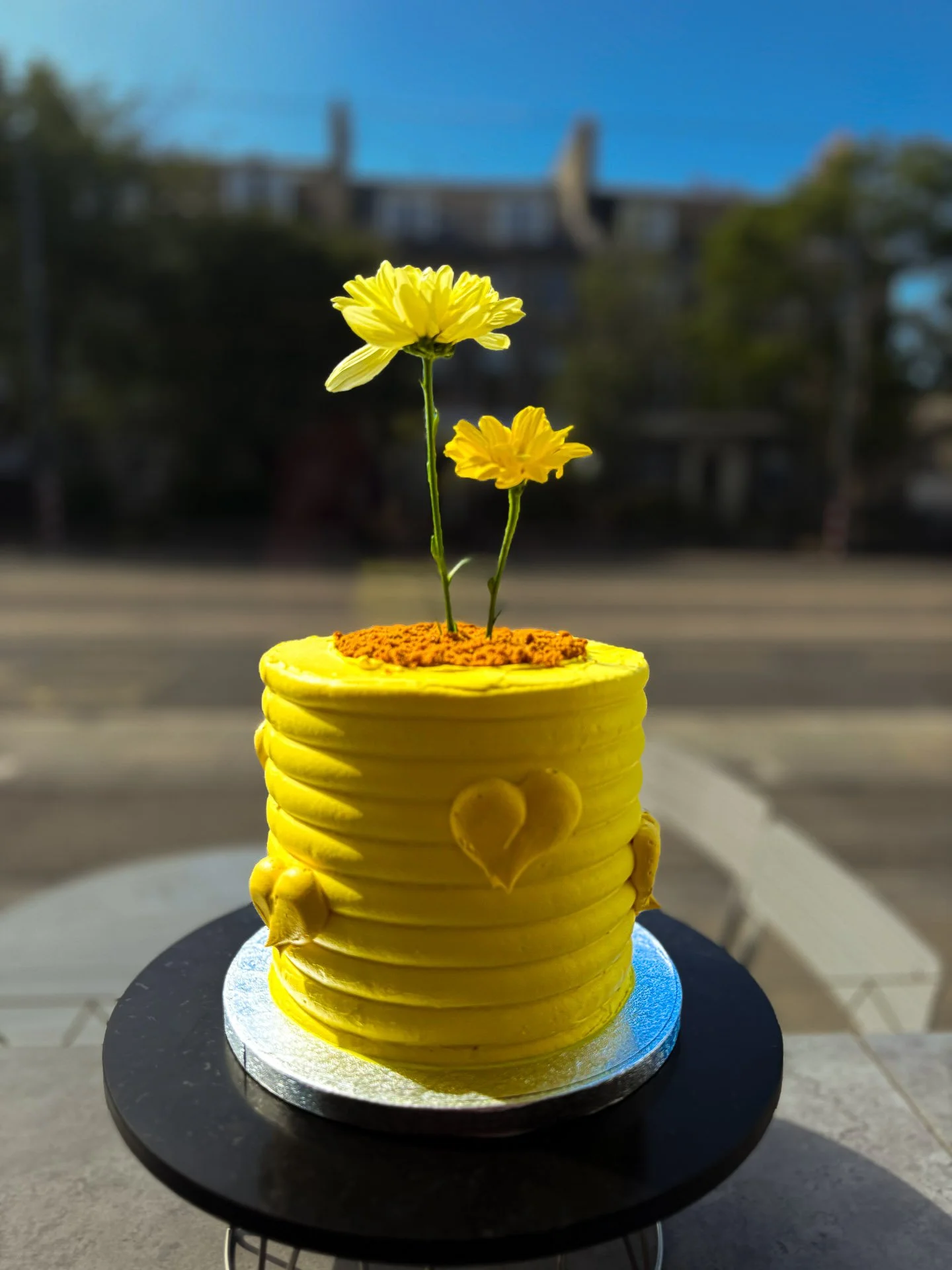 A yellow cake with decorative hearts, topped with two yellow flowers growing from orange sprinkles, placed on a cake stand outdoors with a blurred background of buildings and trees.