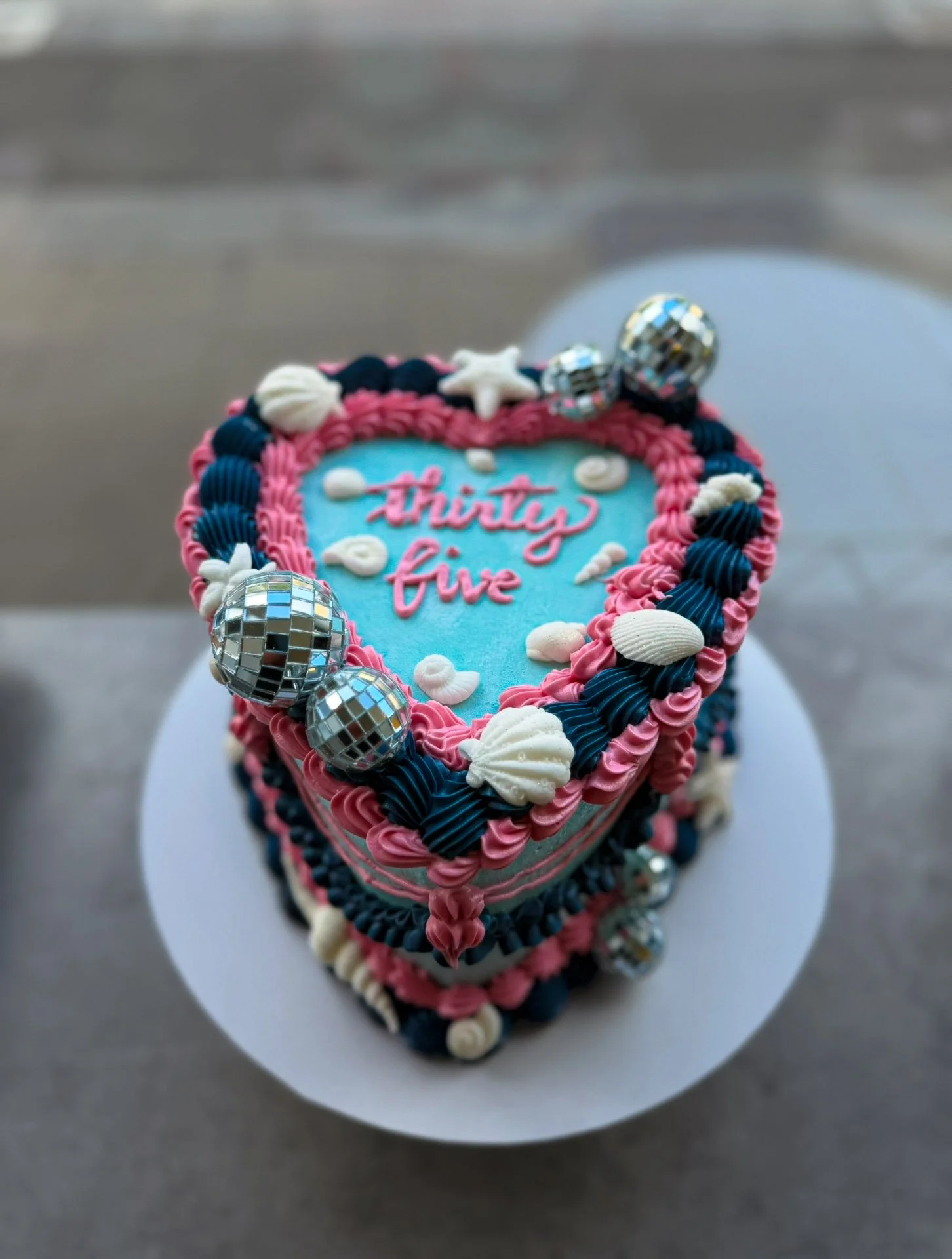 Heart-shaped birthday cake decorated with blue, pink, and white icing, silver disco ball decorations, and edible shells. The cake has the words 'Thirty five' written on top.