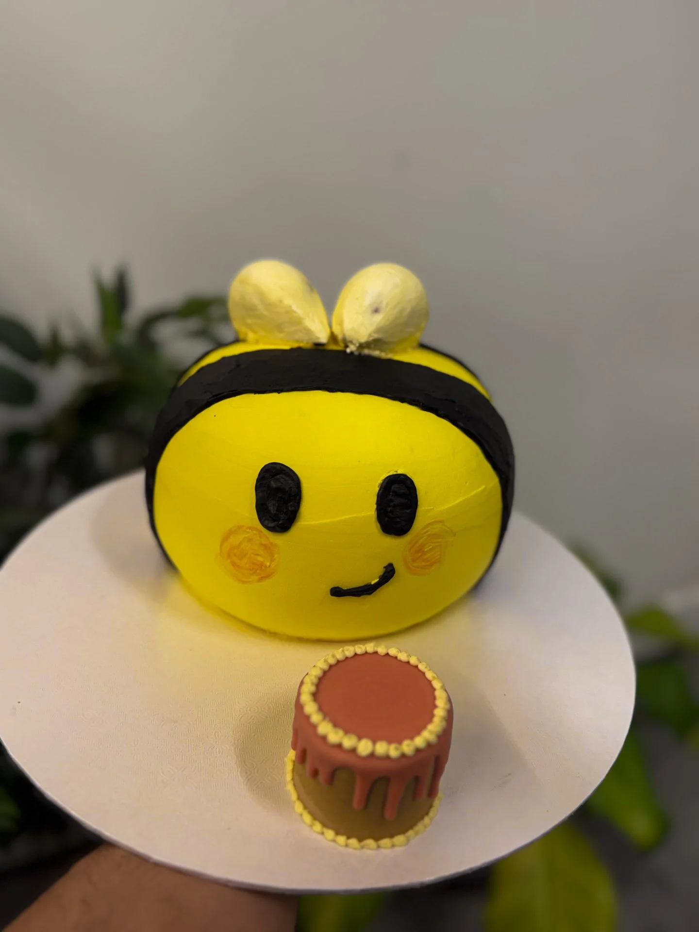 Decorative yellow bee-shaped object with black stripes, a happy face, and a small cake on a white plate.