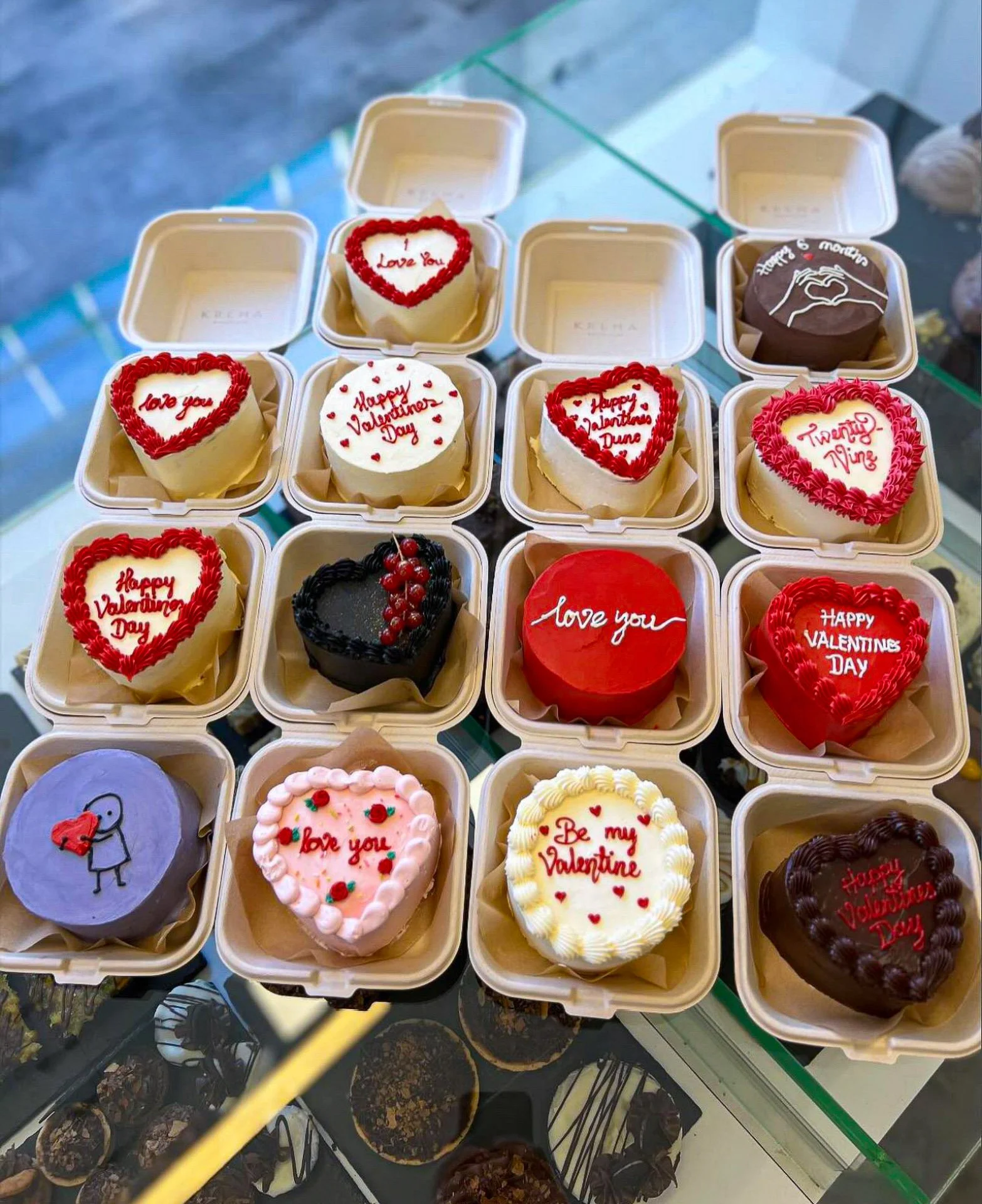 An assortment of decorated Valentine's Day and love-themed small cakes in white boxes on display, with various messages like 'Love You' and 'Happy Valentine's Day,' some with heart decorations and others with illustrations, arranged in three rows wit