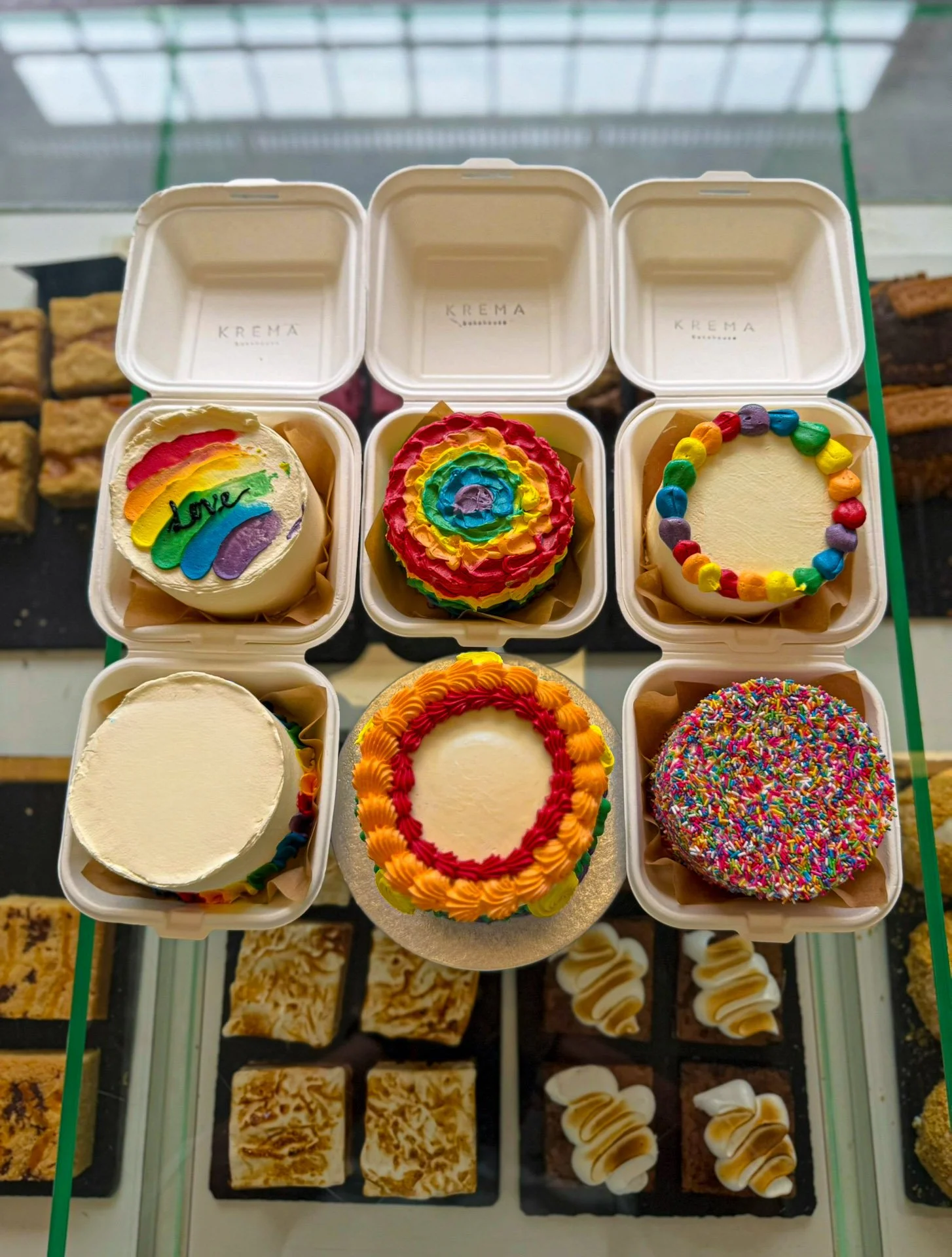 Six cupcakes with colorful rainbow, swirl, and confetti decorations sit in open boxes on a glass display case.
