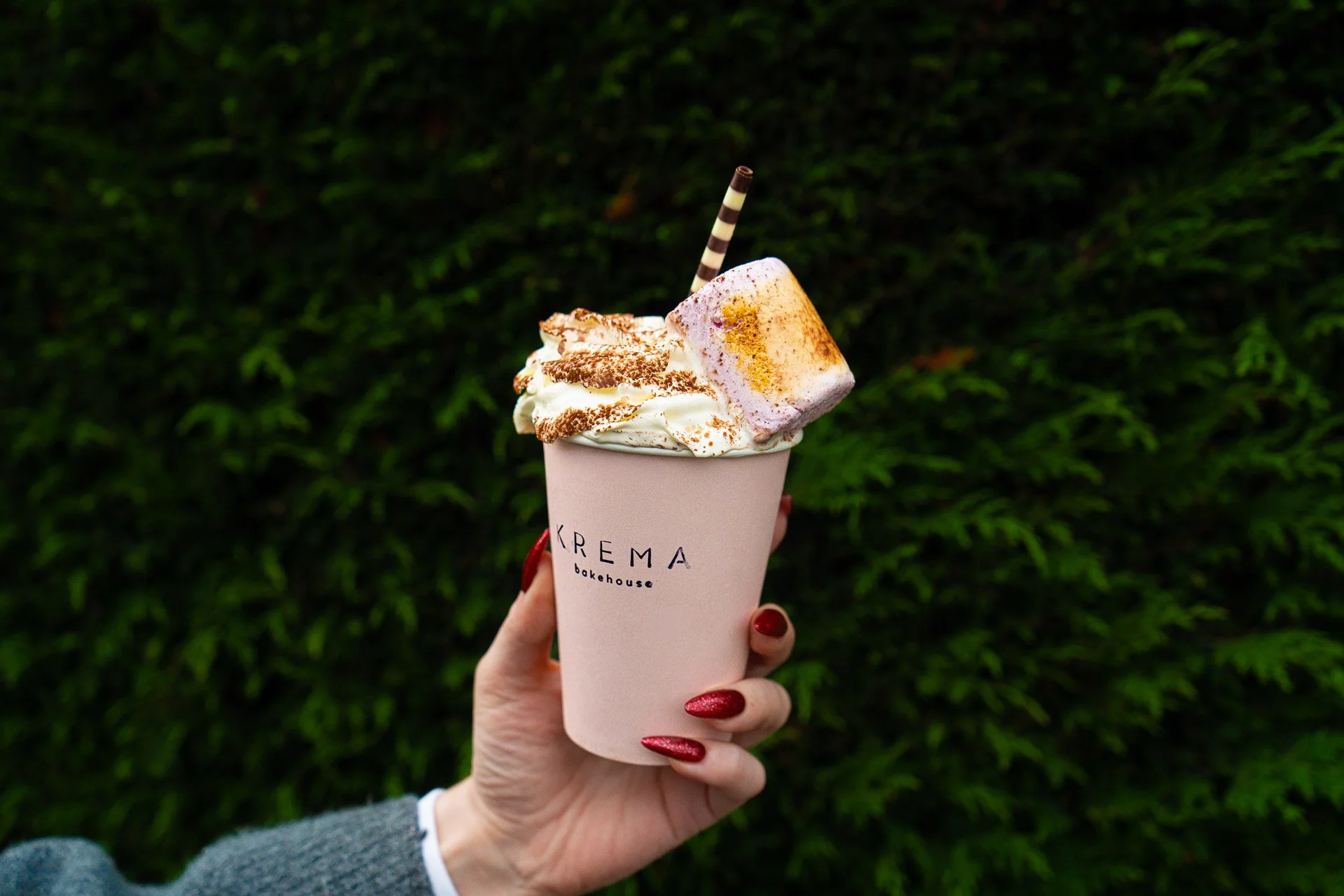 A hand with red manicured nails holding a cup from KREMA bakehouse, topped with whipped cream, chocolate shavings, a toasted marshmallow, and a striped straw, against a background of green foliage.