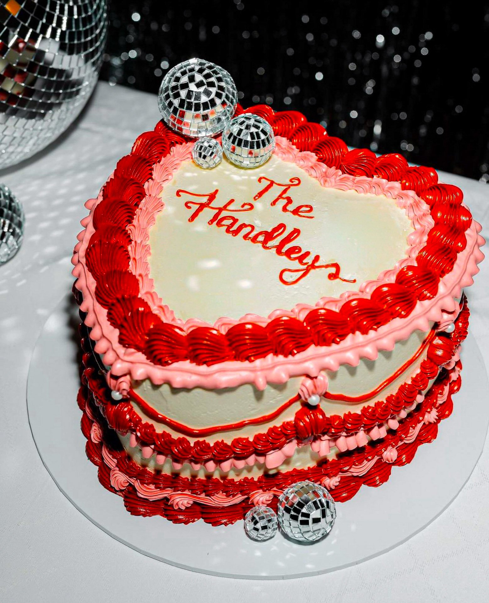 Heart-shaped cake with pink and red frosting, silver disco balls on top, and red cursive writing that says 'The Handley's'.