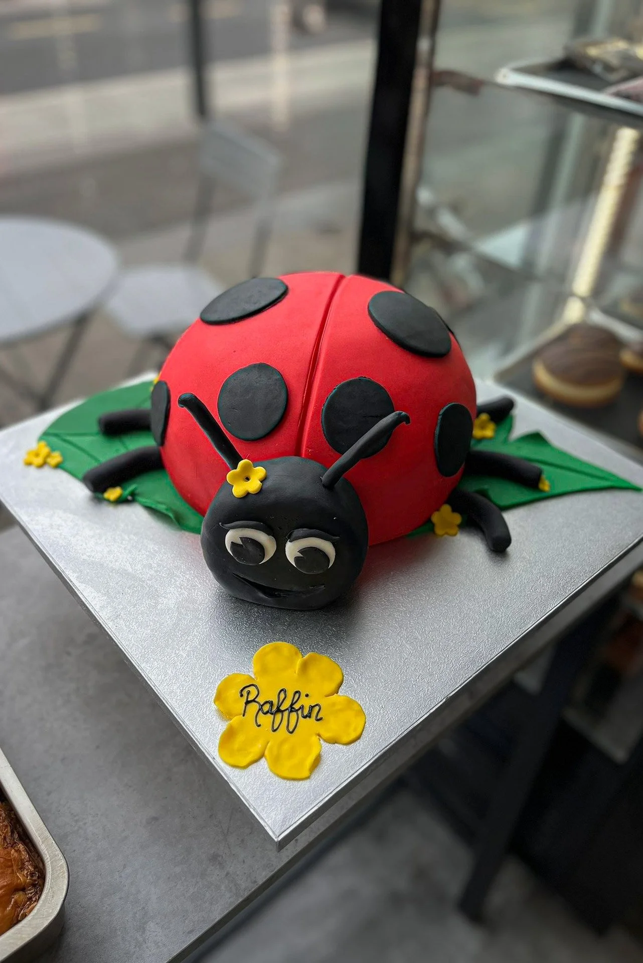 Ladybug-shaped cake with red body, black spots, and a smiling black head with white eyes, placed on a silver-colored board with a yellow flower and the name 'Raffin' written on it.
