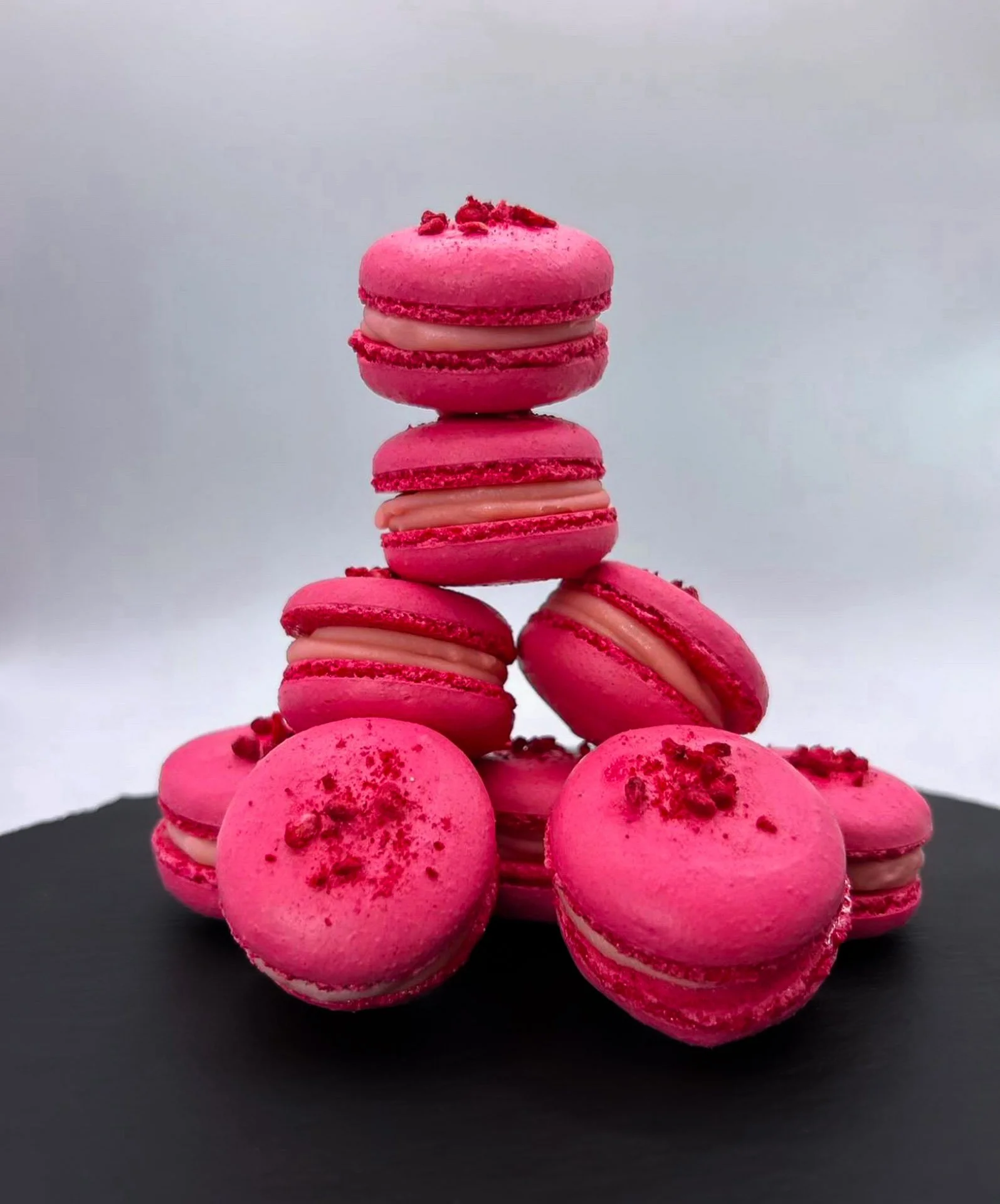 Pink macarons stacked in a pyramid, with some macarons on a black surface in the foreground and a plain white background.