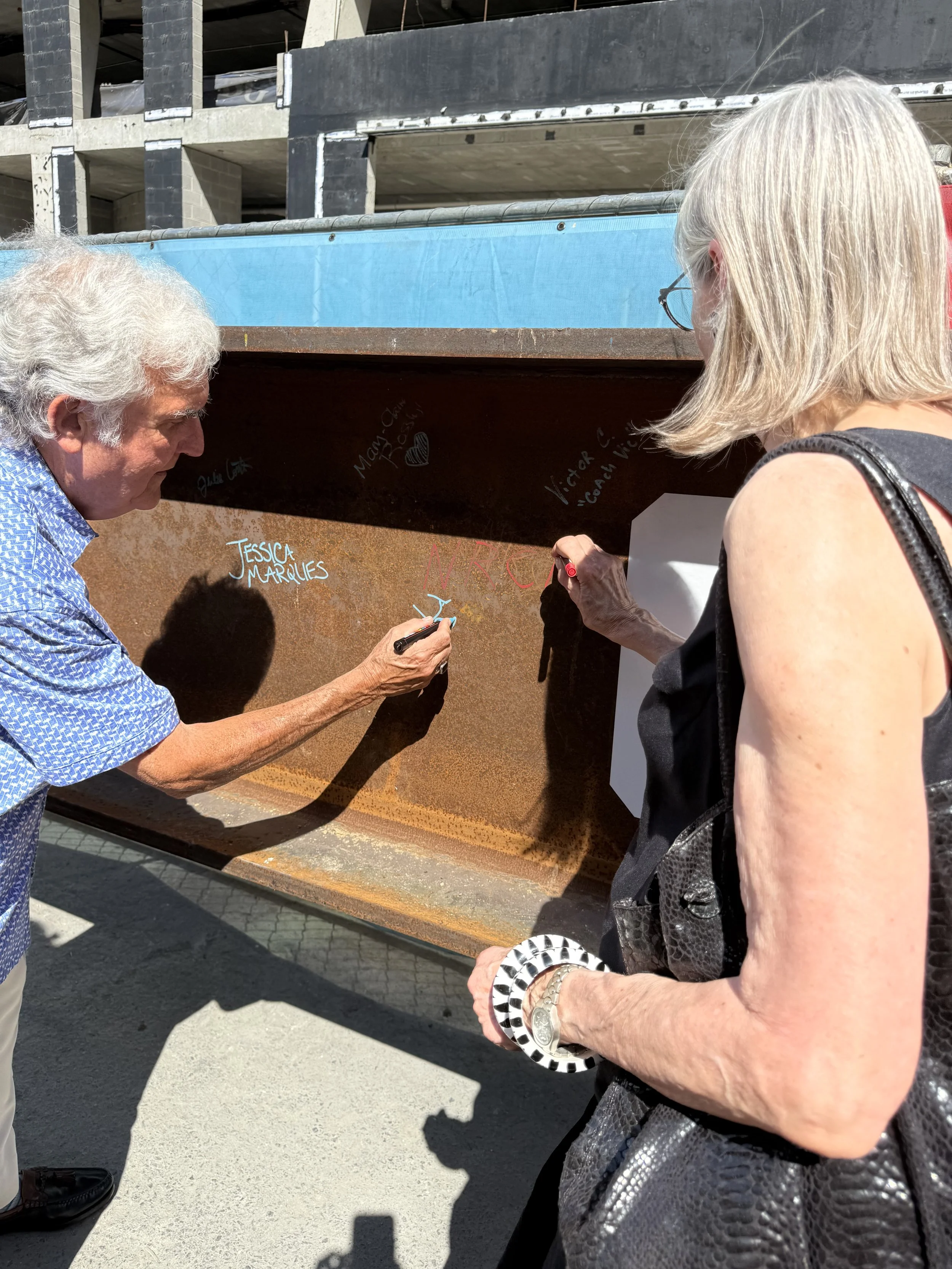 An elderly man with white hair and a blue patterned shirt is writing on a rusted metal surface with a black marker. A woman with blonde hair, glasses, and a black sleeveless top is observing and holding a black and white checkered object. The scene t