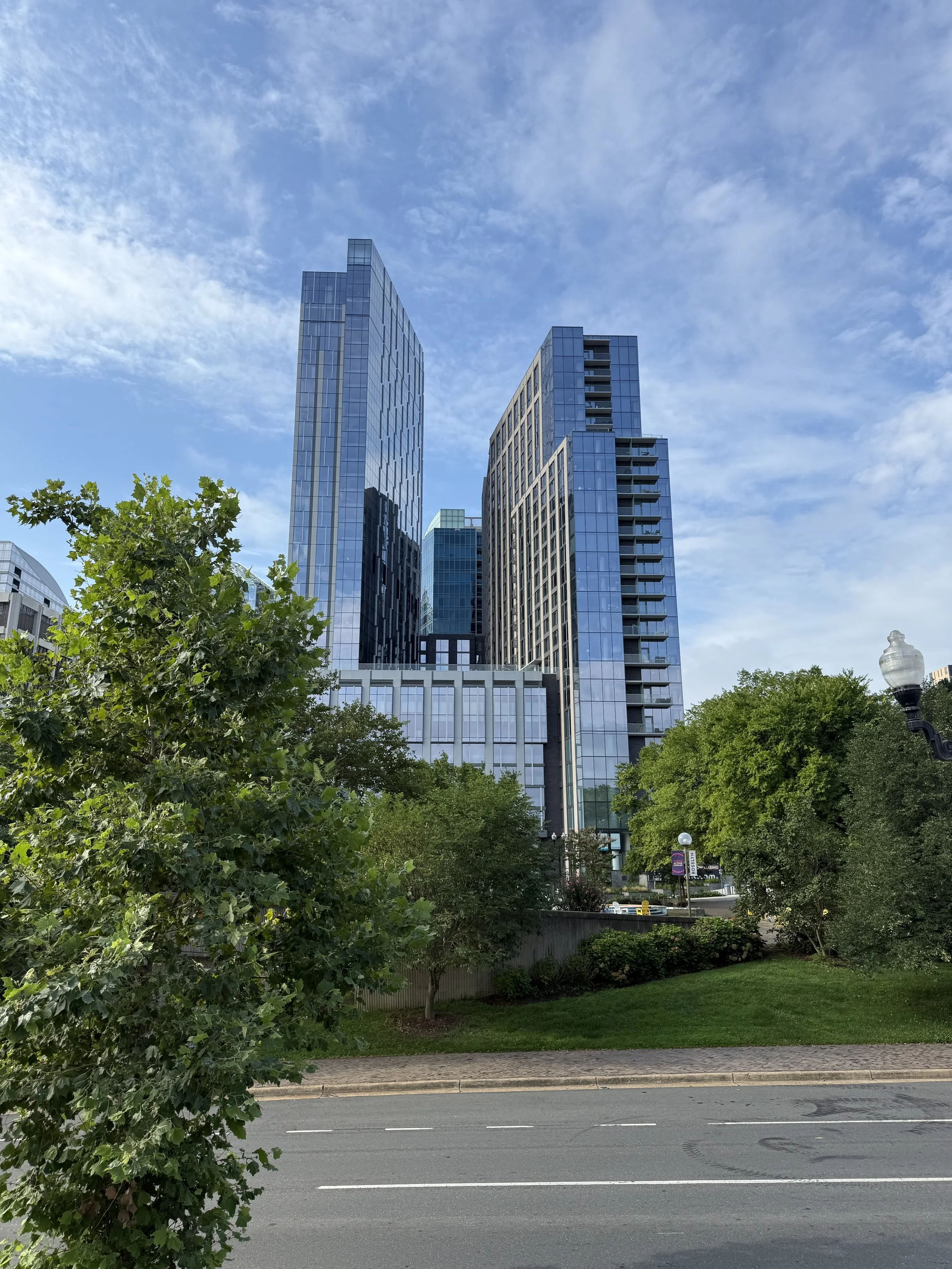 Tall modern glass buildings with a blue sky and white clouds, green trees and a sidewalk in the foreground.