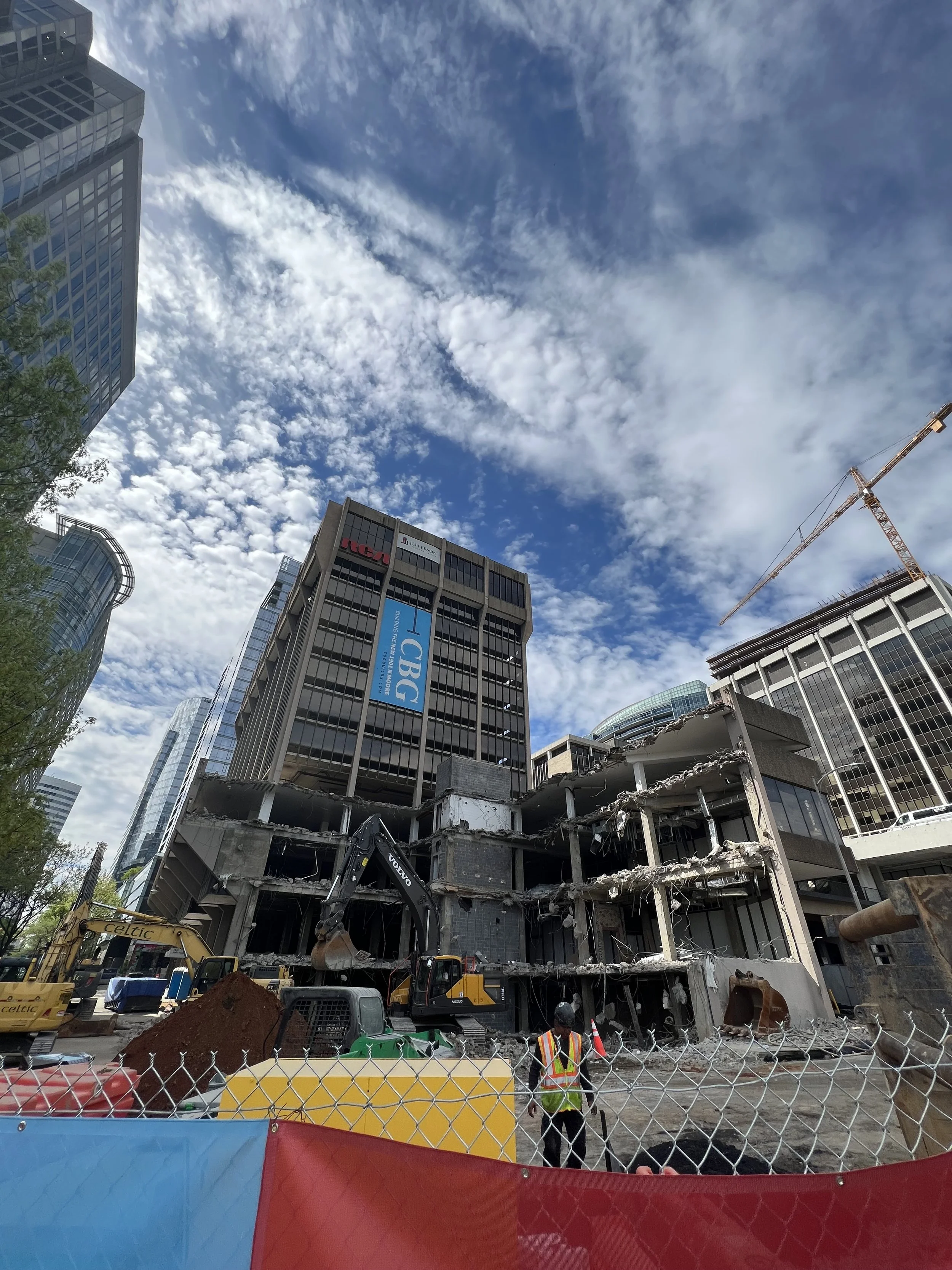 Construction site with a partially demolished building, construction machinery, and workers in safety vests, surrounded by tall office buildings and a blue sky with scattered clouds.