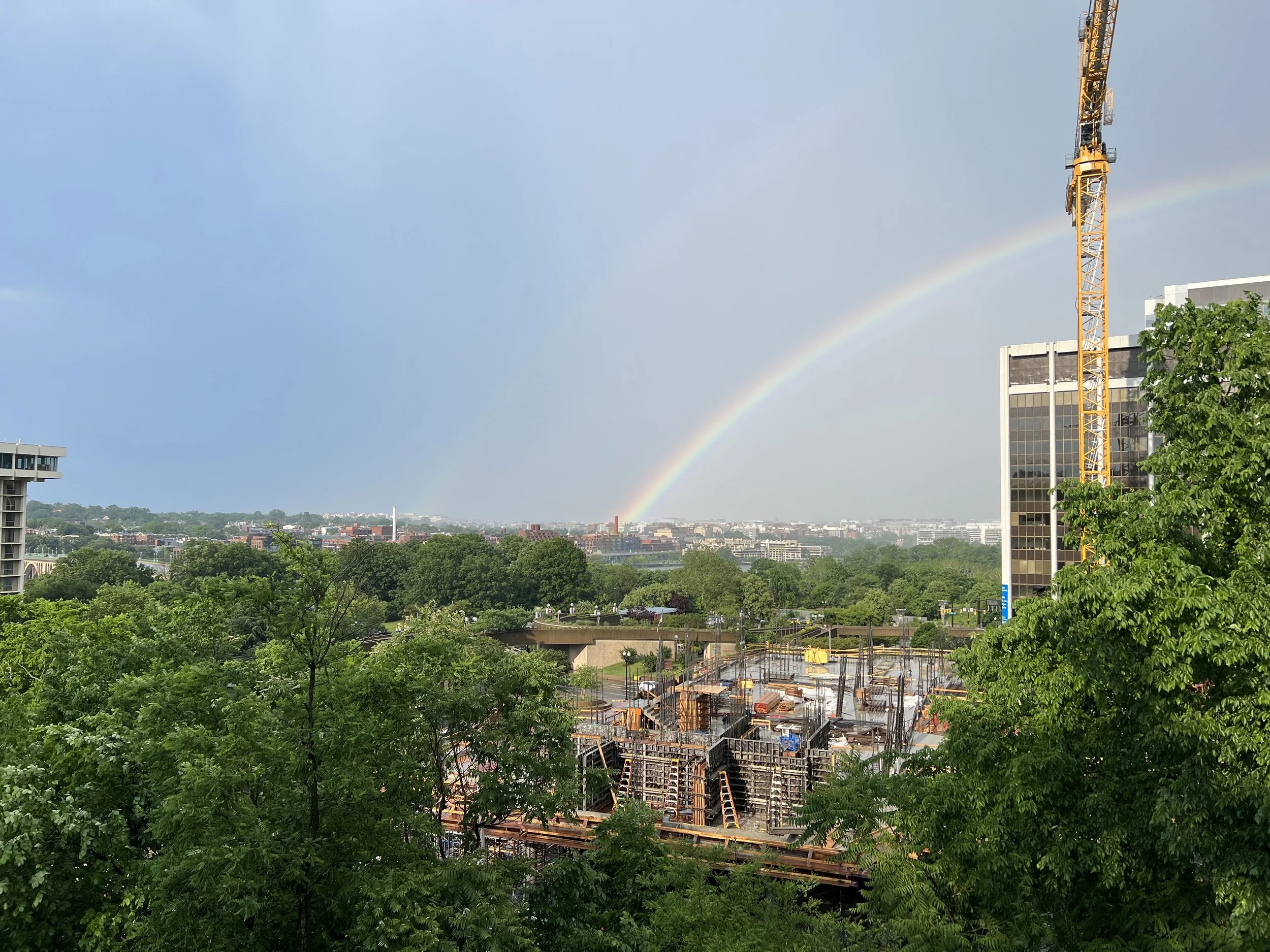A cityscape with green trees in the foreground, a construction site with steel structures, a tall yellow crane, and a rainbow in the sky over buildings.
