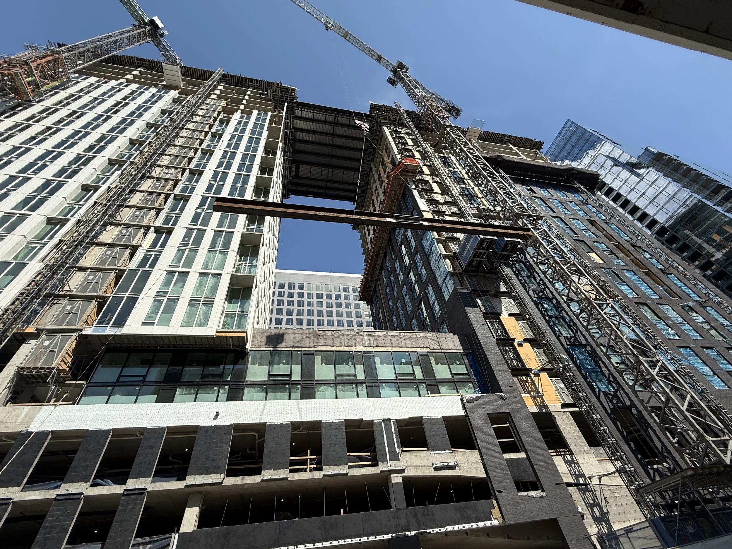 Construction site of a high-rise building with cranes, scaffolding, and partially completed glass windows, under a clear blue sky.